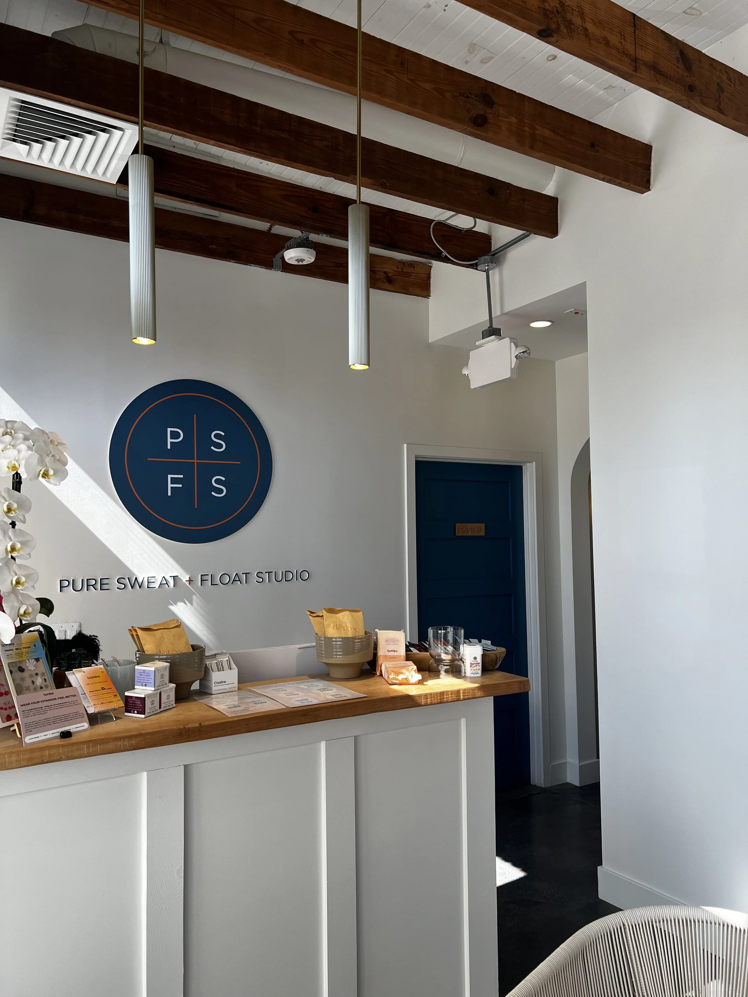 Reception area of Pure Sweat + Float Studio with a counter displaying products, a wall sign with the studio's logo, and a doorway leading to another room.