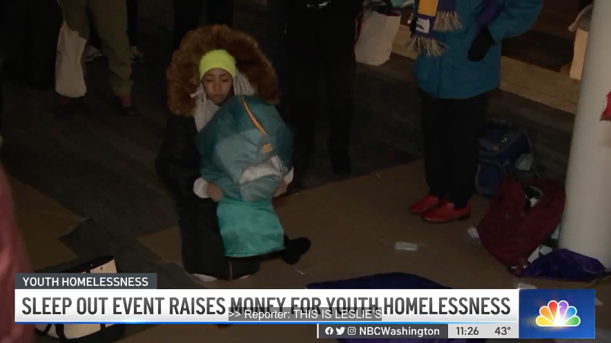 A young girl with curly hair wearing a yellow headband, a black winter coat, and gloves, hugging a sleeping bag with another person inside it during a sleep out event for youth homelessness.