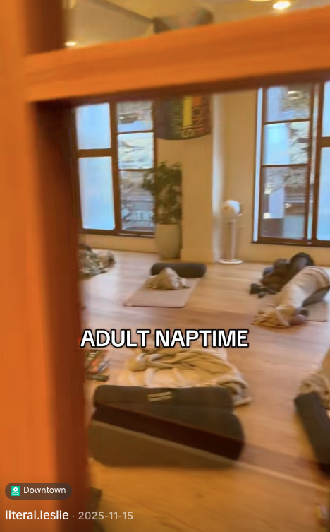 A cozy room with large windows, wooden floors, and various sleeping bags and cushions on the floor, labeled 'Adult Naptime'.