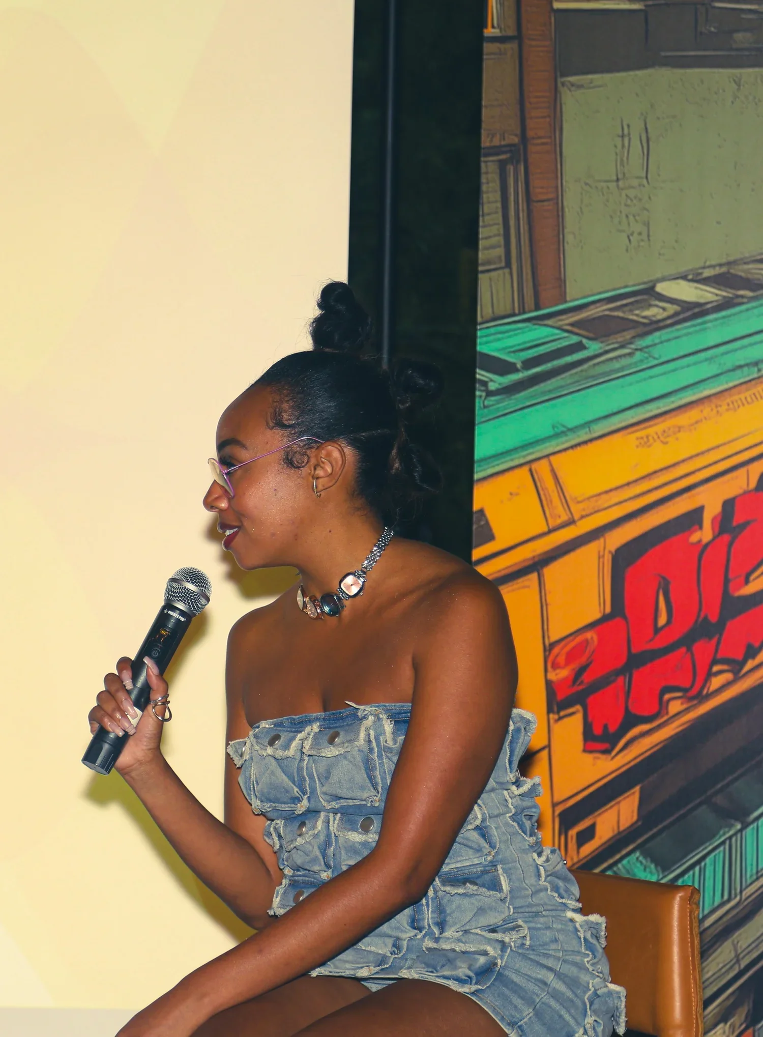 A woman with dark hair styled in buns is sitting and speaking into a microphone. She is wearing glasses, a necklace, and a distressed denim tube top. Behind her, there is a colorful poster or mural featuring text and illustrations.