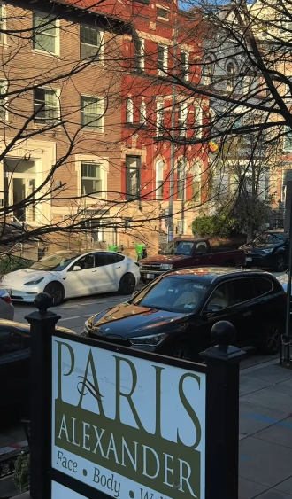 Street view with parked cars and buildings in the background. A sign in the foreground reads "PARIS ALEXANDER Face Body W".