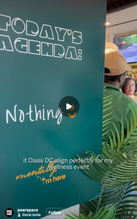 A teal-colored sign with white and yellow text in front of a woman and a man at a wellness event, with plants and indoor decor in the background.