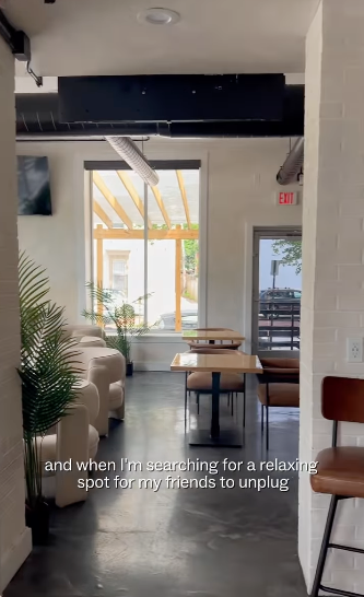 Interior of a modern cafe with white walls, natural light, a potted plant, and seating including a white armchair and wooden tables with chairs.