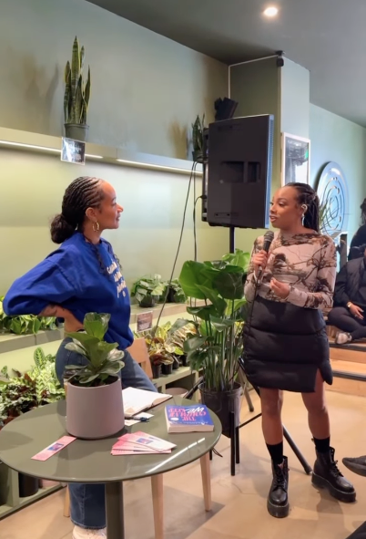Two women speaking at an event near a table with potted plants and pamphlets, with a speaker and audience in the background.