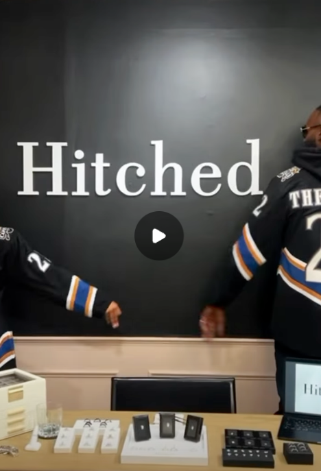 Two people wearing hockey jerseys interacting in front of a wall with the word "Hitched" on it. There is a table with small boxed items, a laptop, and other objects in front of them.