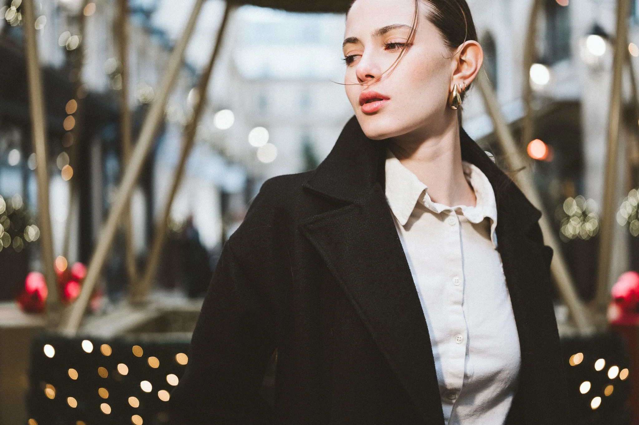 A woman wearing a black coat and white shirt standing at the village royale in a Paris outdoor shopping area with holiday decorations and blurred lights in the background.