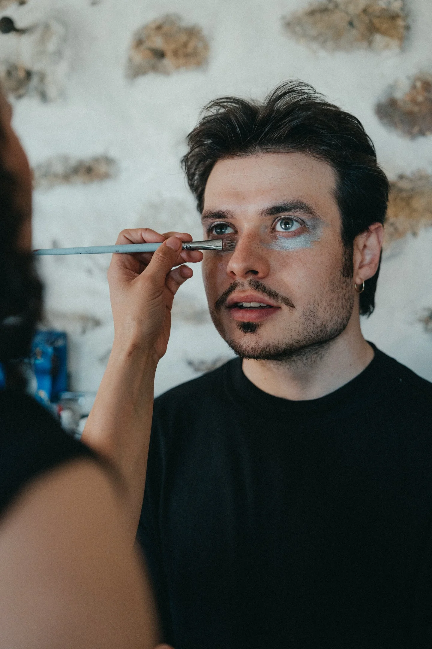 A man with short dark hair, blue eyes, and facial hair, lying back while makeup artist applies makeup with a brush under his eye.