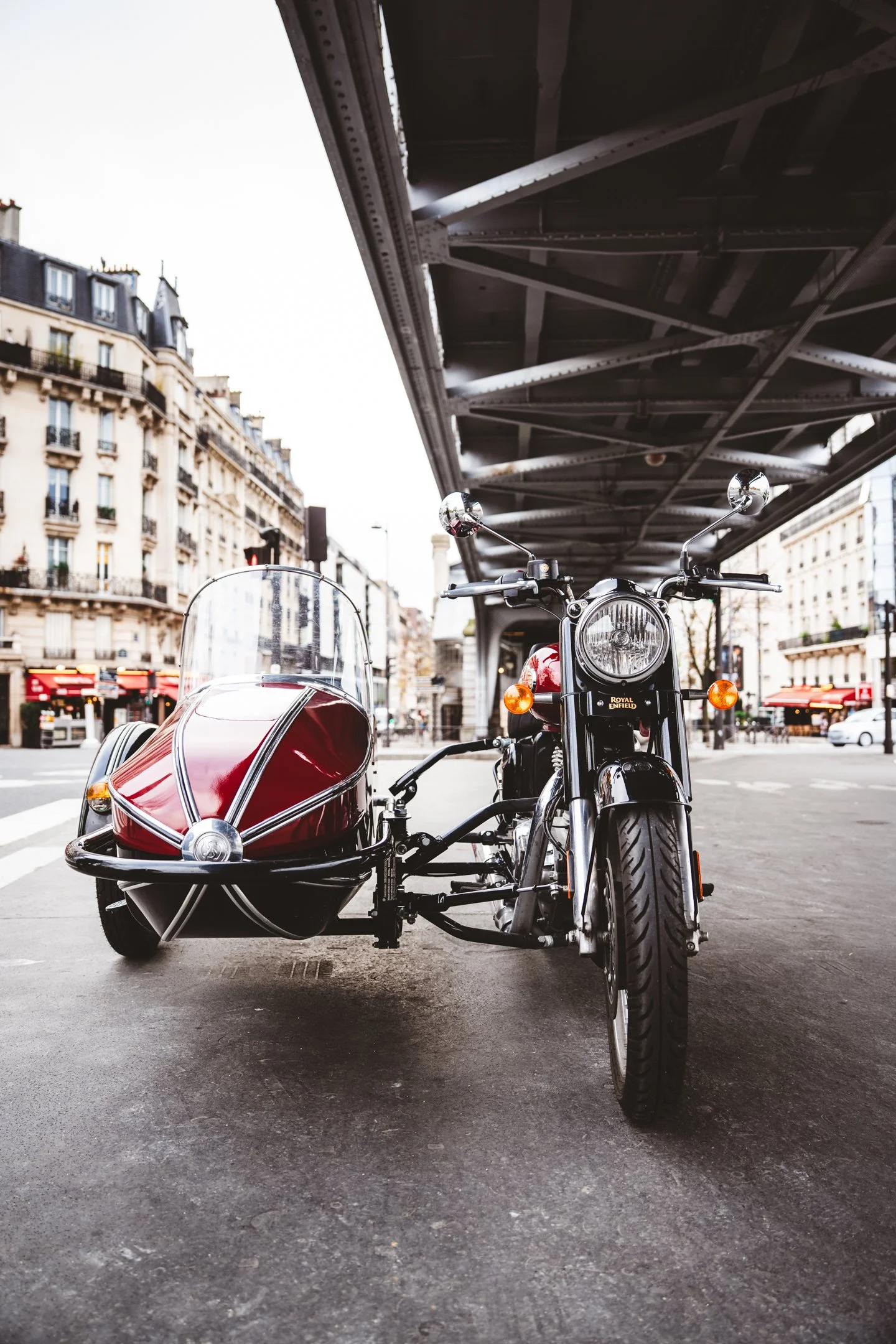 A vintage motorcycle with a sidecar parked under a bridge in a city street. The motorcycle is black and chrome with a Round Enfield logo on the front. The sidecar has a red and white striped pattern. Buildings with classic European architecture line 
