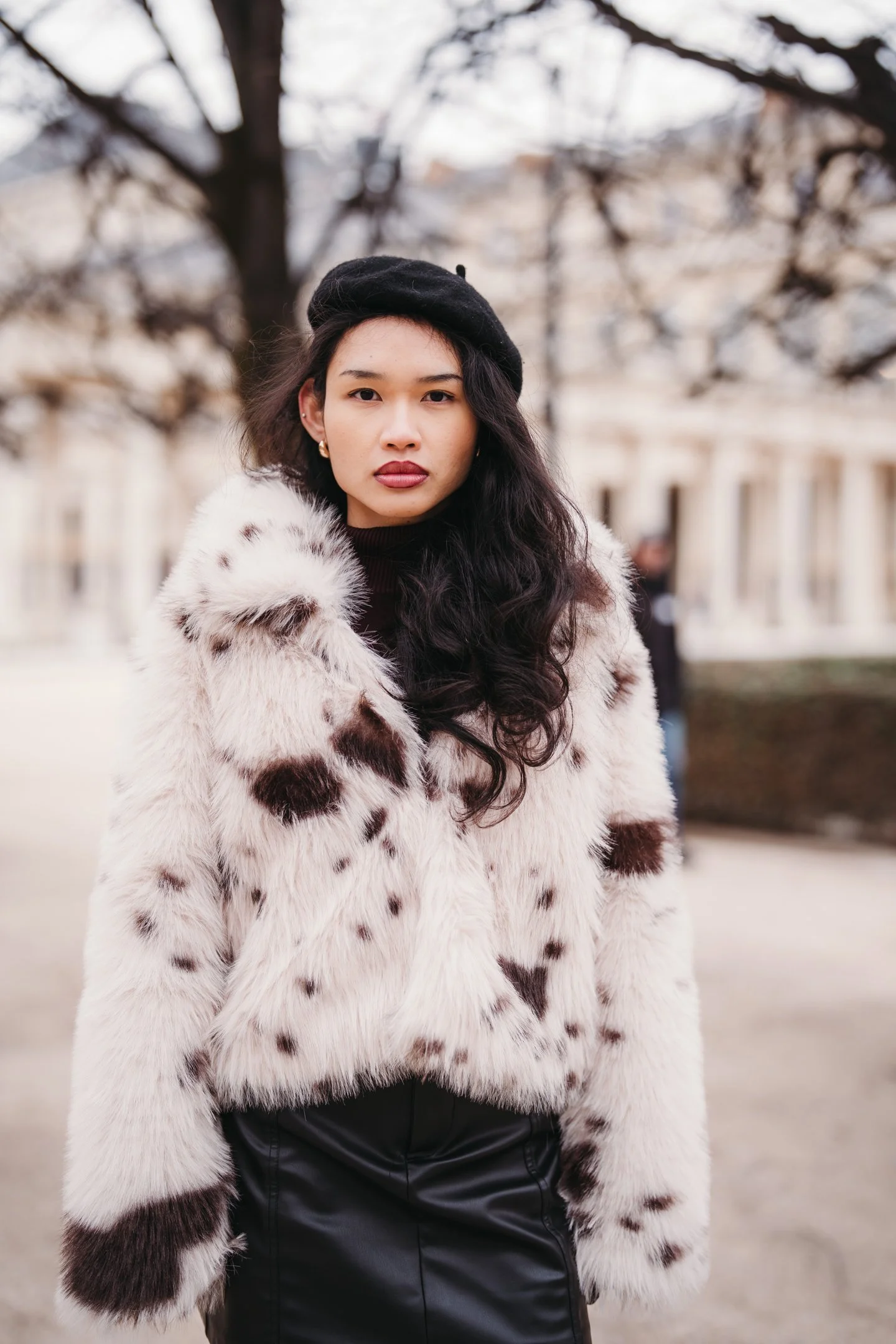A woman with long dark wavy hair wearing a black beret and a cream and brown animal print fur coat, standing outdoors with blurred trees and buildings in the background.