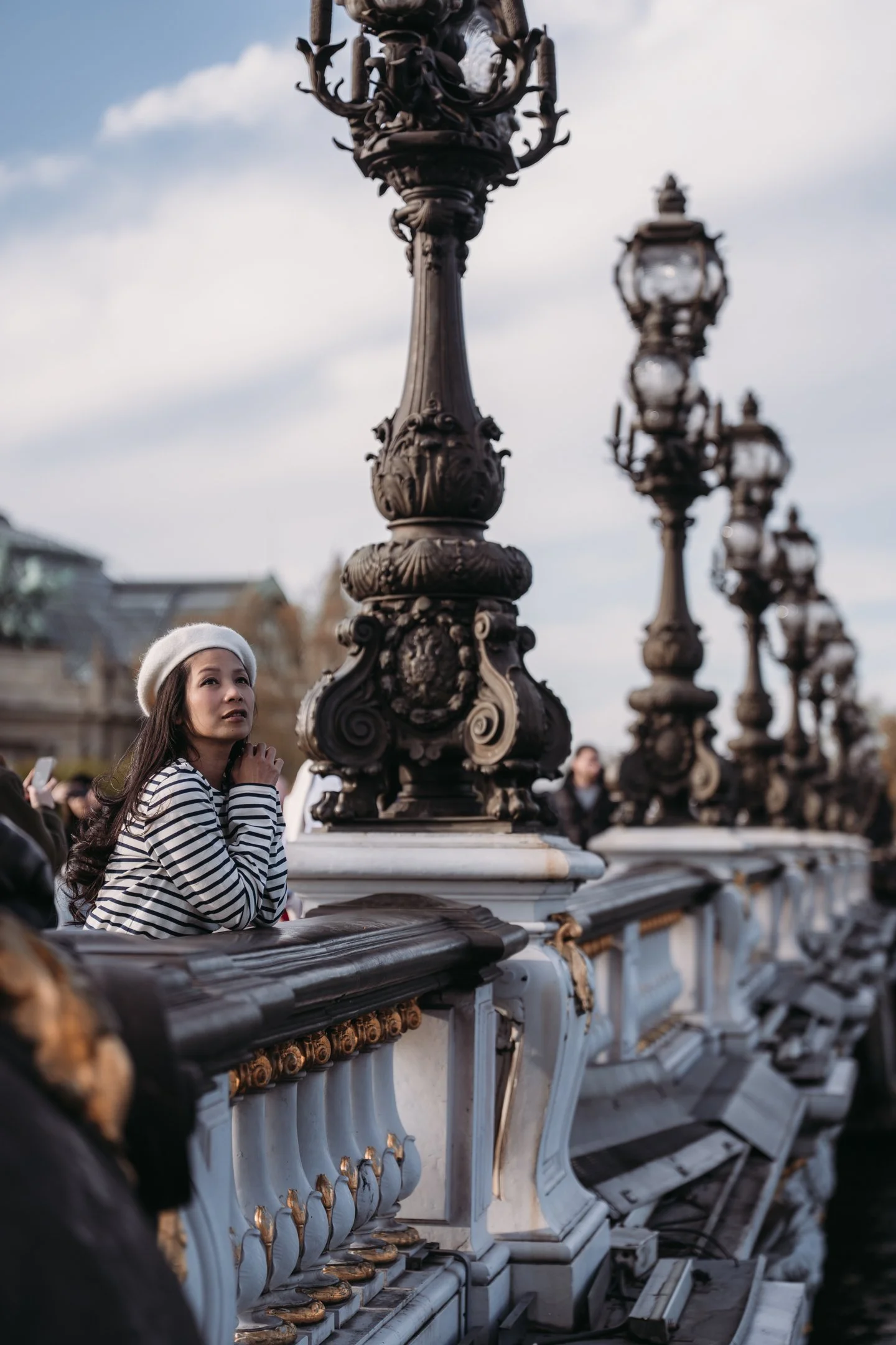 A woman wearing a white beret and a black-and-white striped shirt standing on a bridge, looking up, surrounded by ornate street lamps, with a cloudy sky in the background.