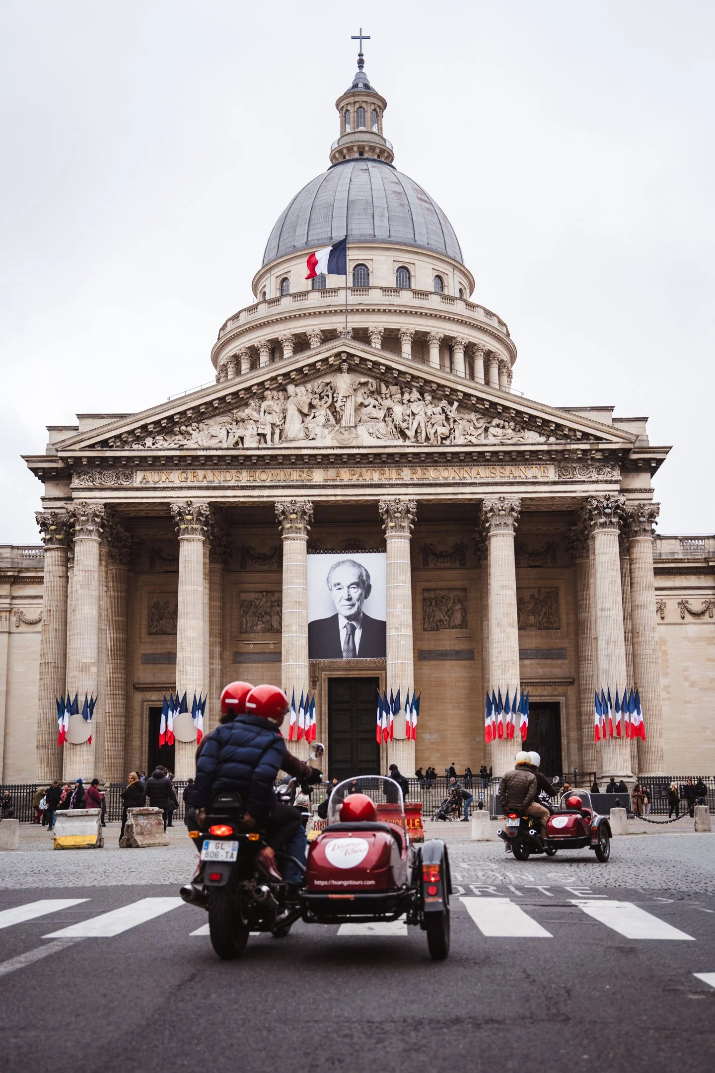 The image shows the front of the Panthéon in Paris, France, decorated with French flags and a large portrait of a man. People are riding scooters and walking in front of the building on a cloudy day.