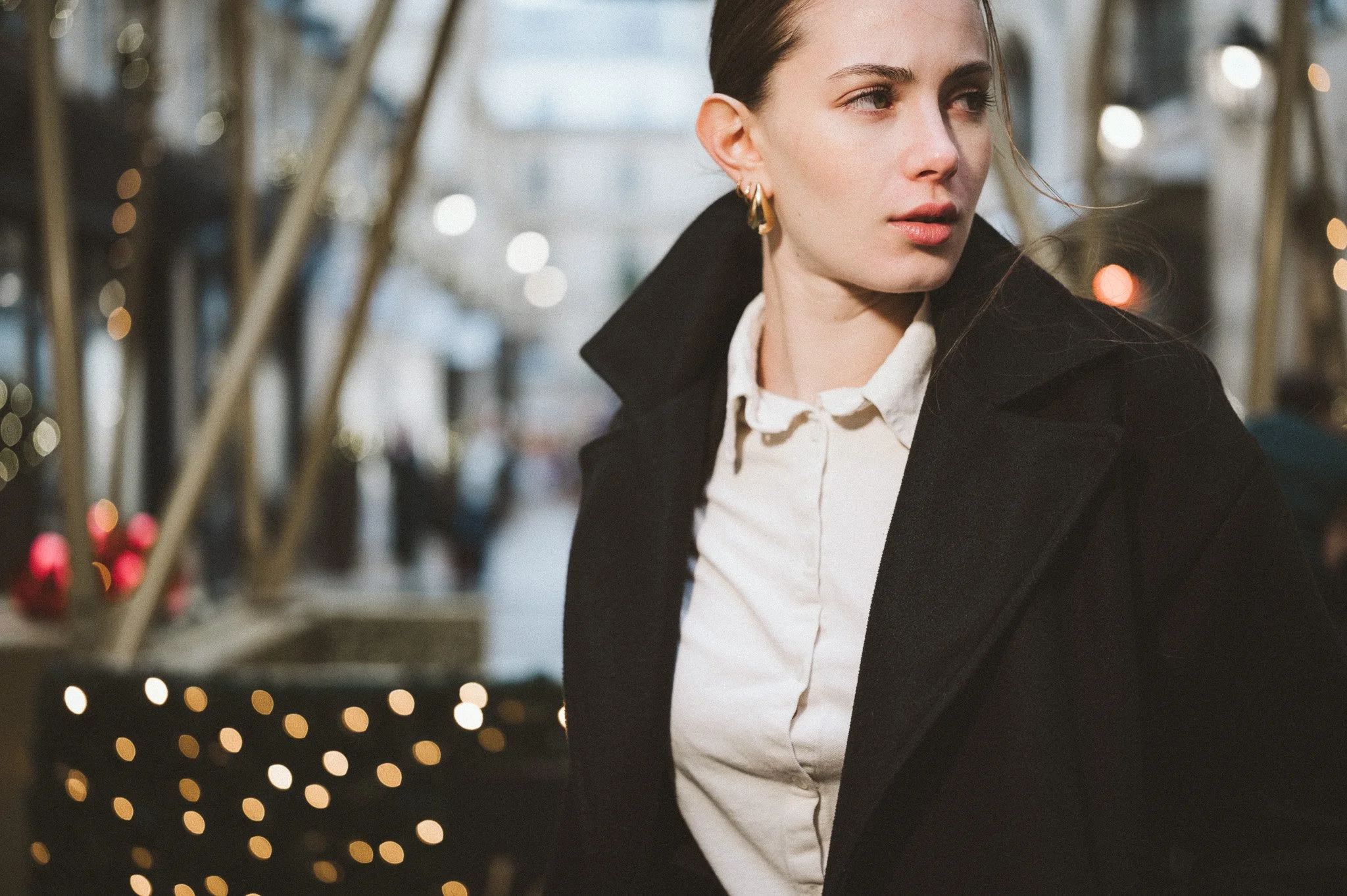 A woman wearing a black coat and white shirt standing at the village royale in a Paris outdoor shopping area with holiday decorations and blurred lights in the background.