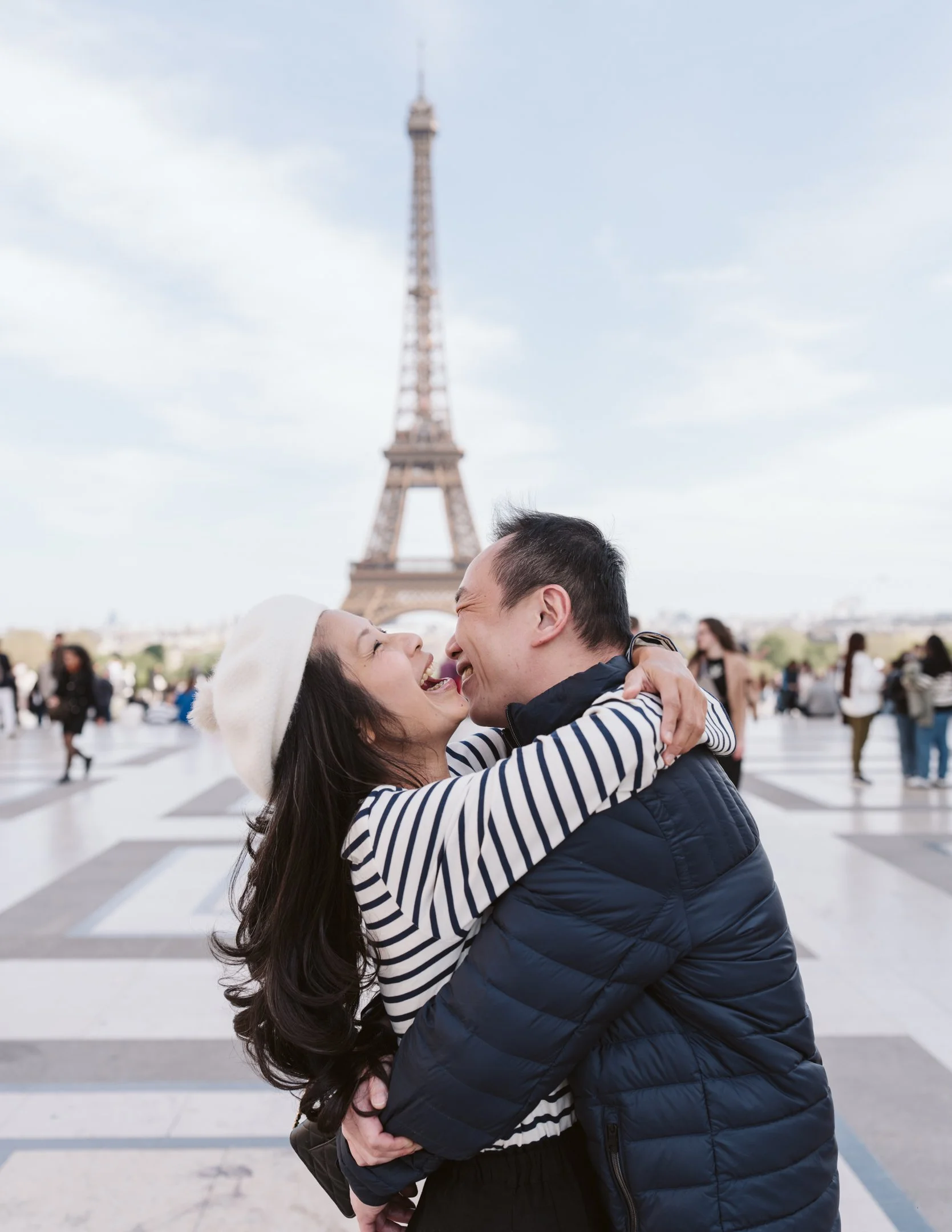 A couple embraces and shares a kiss in front of the Eiffel Tower in Paris, France, with other tourists around them.