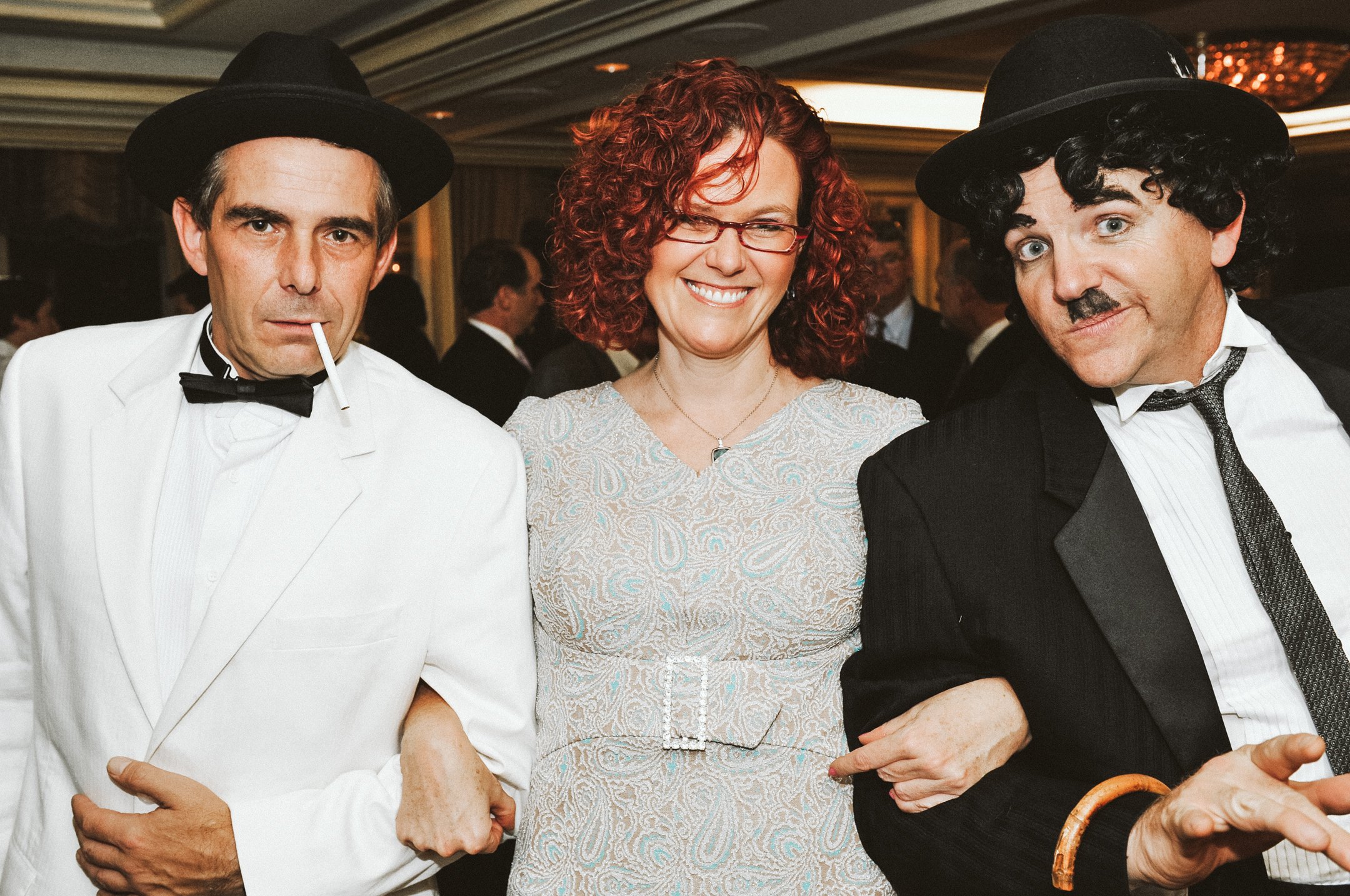 Three people dressed in vintage attire at a social event, with two men in tuxedos and a woman in a light-colored dress, all wearing black hats.