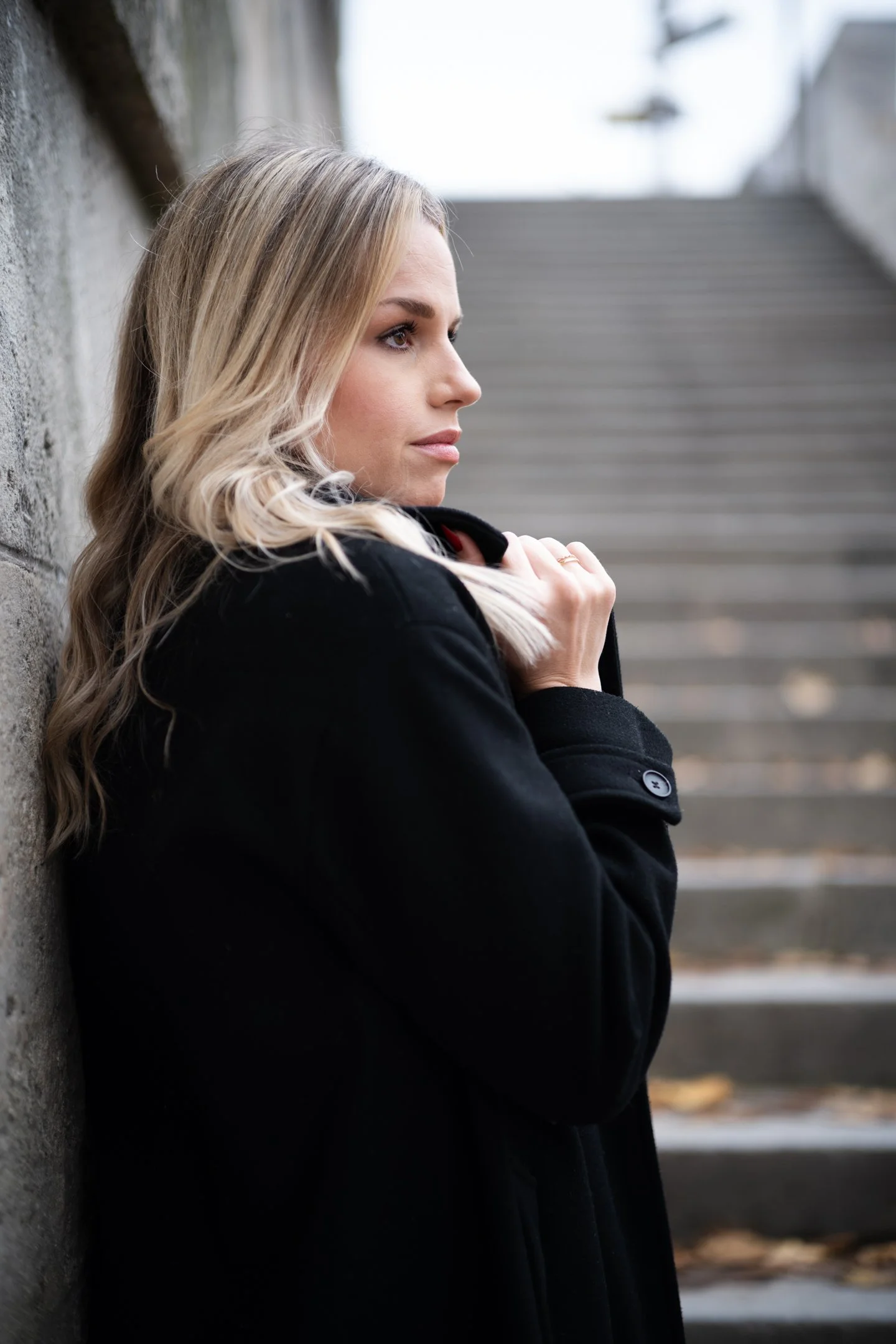 A woman with blonde hair, wearing a black coat, stands against a stone wall outdoors near a staircase, looking to the right with a contemplative expression.