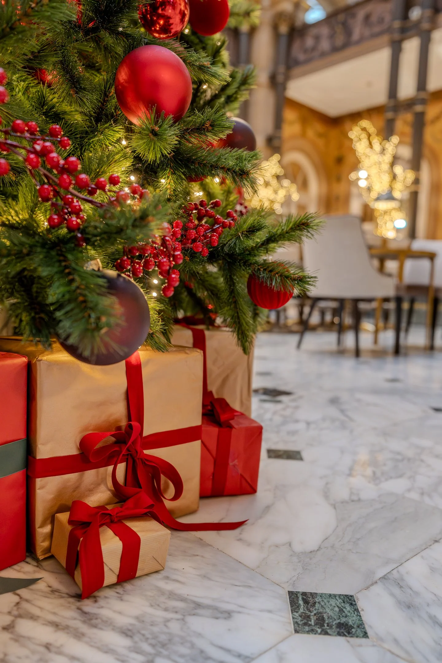 Christmas tree decorated with red and gold ornaments, surrounded by wrapped gift boxes with red ribbons on a marble floor, in an indoor setting with warm lighting and festive decorations.