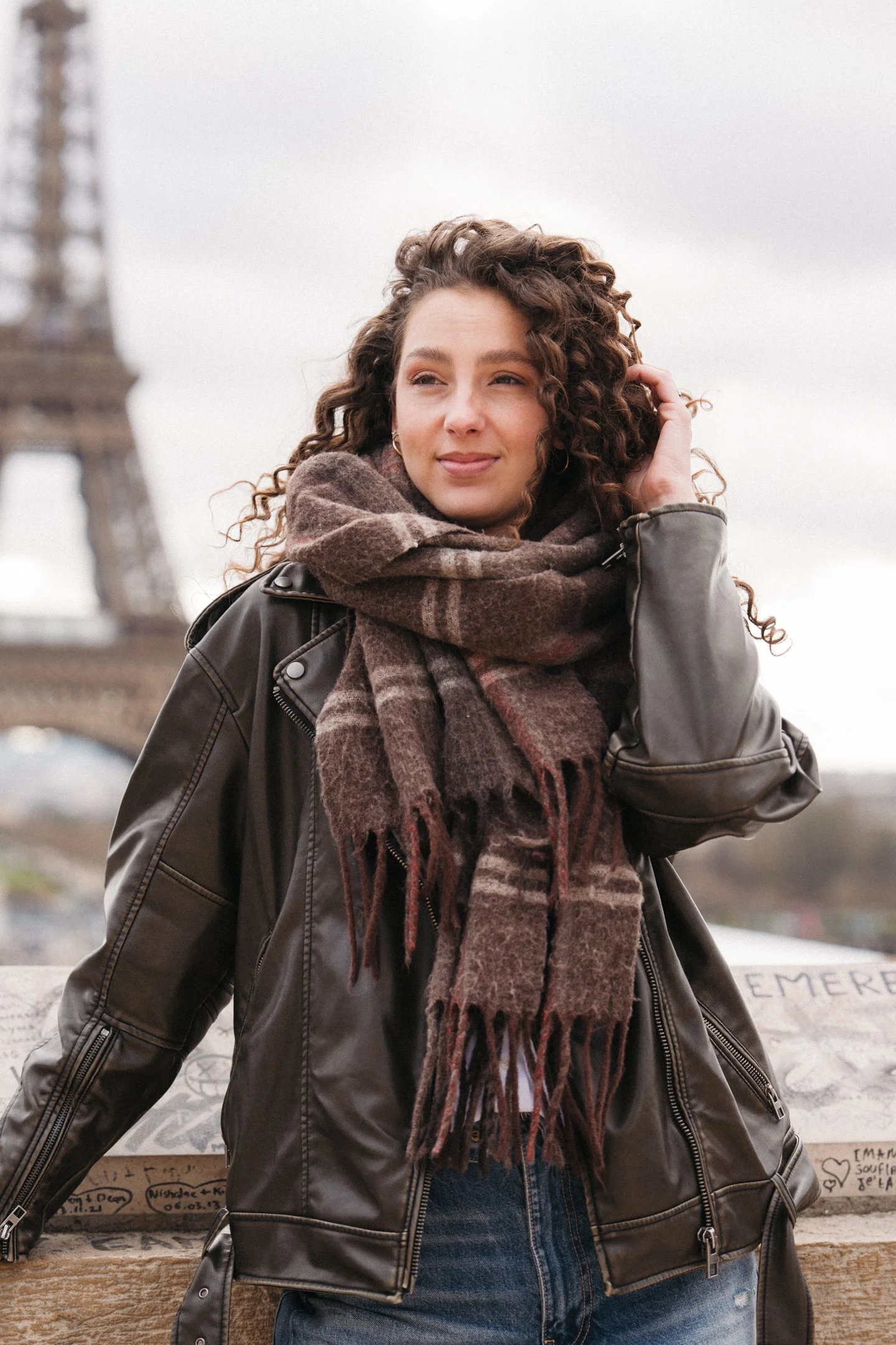 Young woman with curly hair wearing a brown checkered scarf and leather jacket, standing outdoors near a stone memorial with graffiti, with blurred background of a bridge and overcast sky.