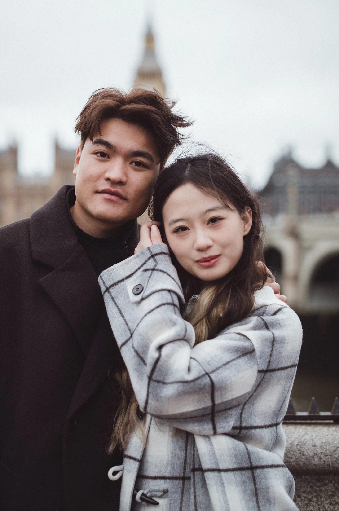 A young man and woman are standing close together outdoors on a cloudy day, with historical buildings and a clock tower in the background. They are embracing and smiling softly at the camera.