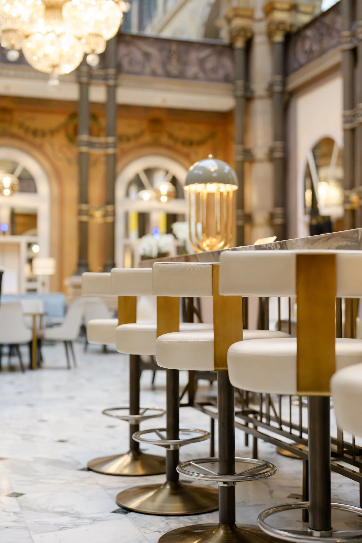 A row of white barstools with wooden backs, lined up along a counter in a stylish, ornate restaurant or cafe with chandeliers and decorative architecture.