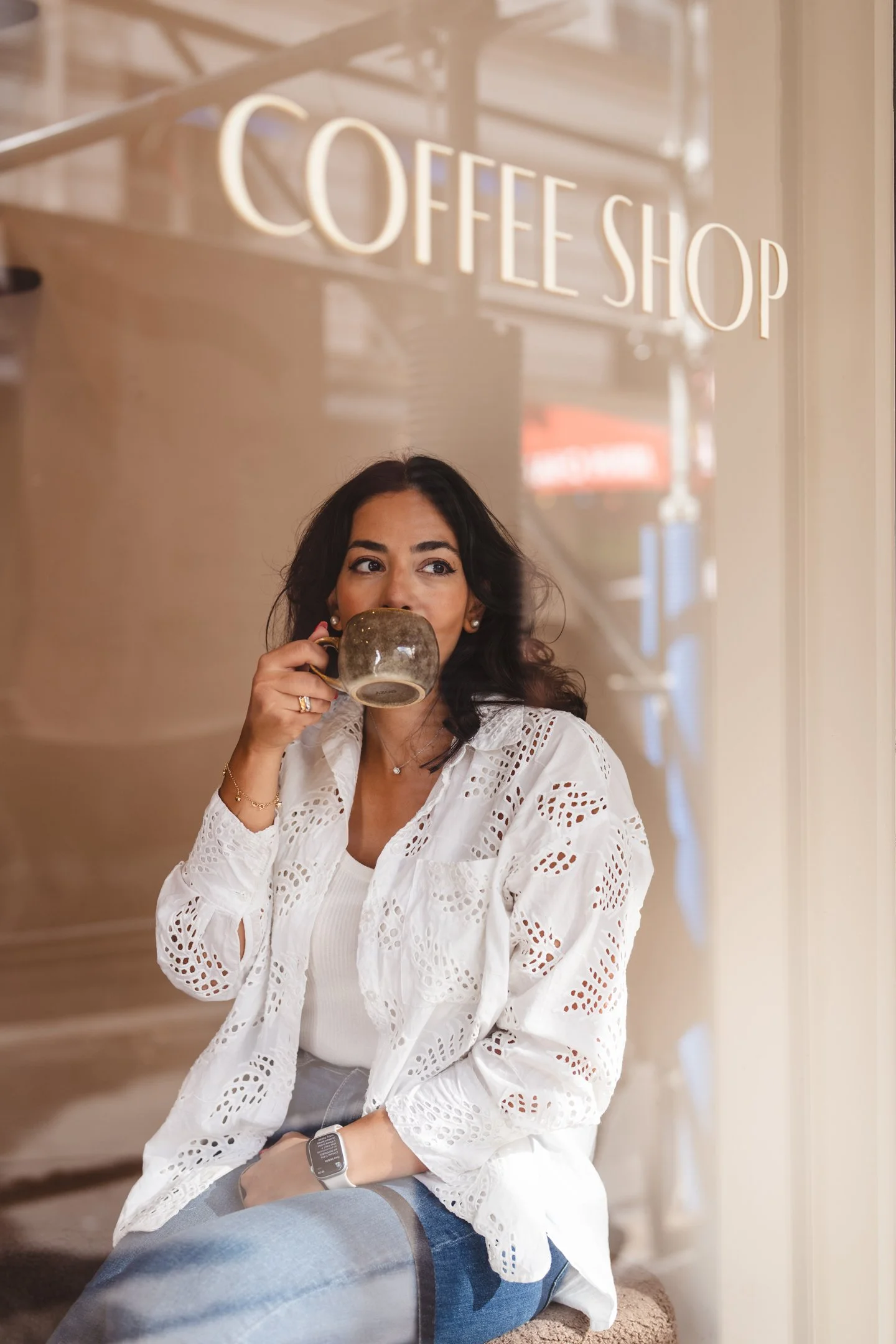 A woman with dark hair drinking coffee inside a coffee shop, wearing a white lace blouse and sitting on a beige cushioned seat.