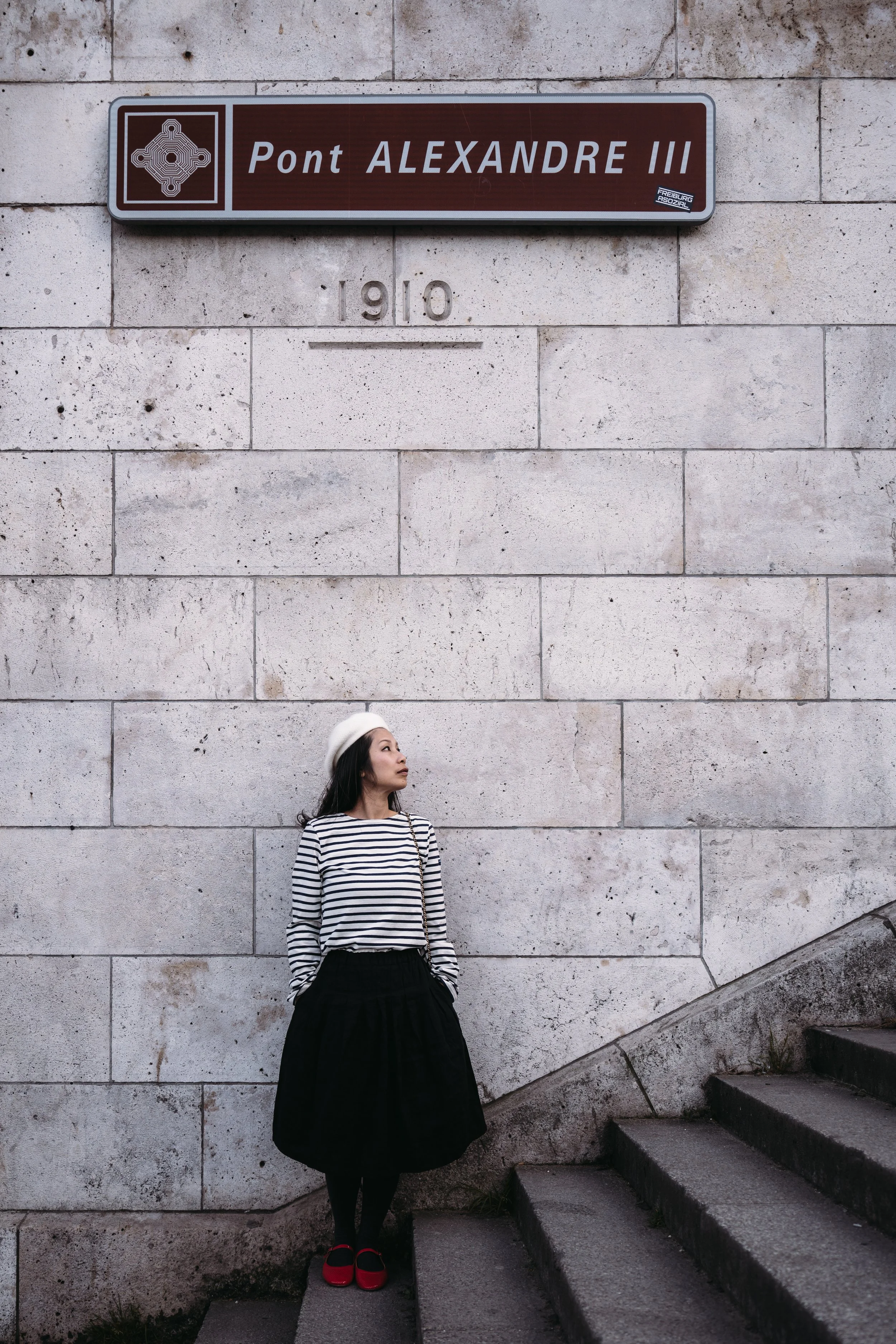 A woman wearing a white beret, striped shirt, black skirt, and red shoes standing against a beige brick wall near steps, with a street sign reading 'Pont Alexandre III' above her.