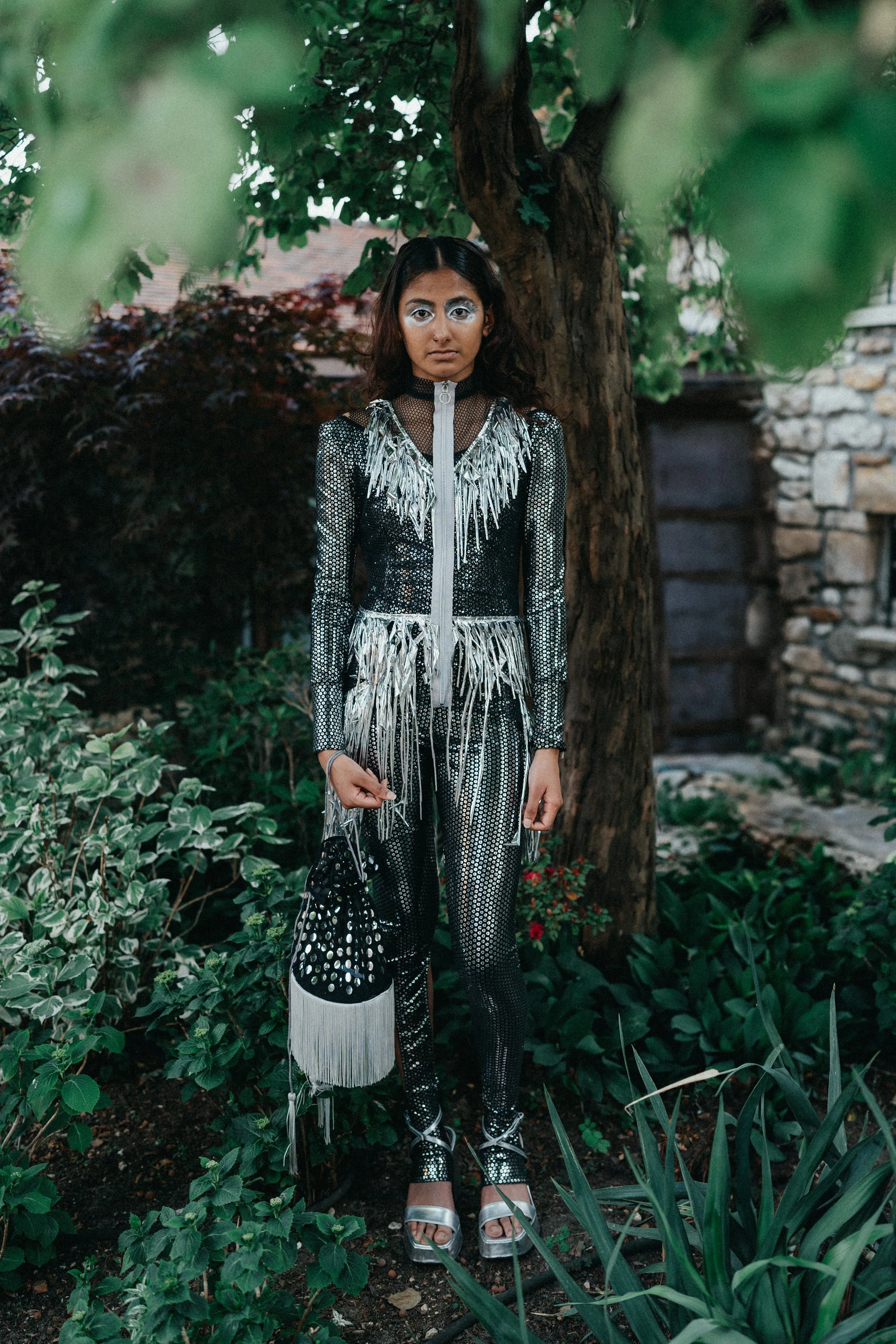 Person standing outdoors in a garden, wearing a shiny black and silver outfit with fringes, holding a black bag with white dots, and wearing high-heeled sandals.