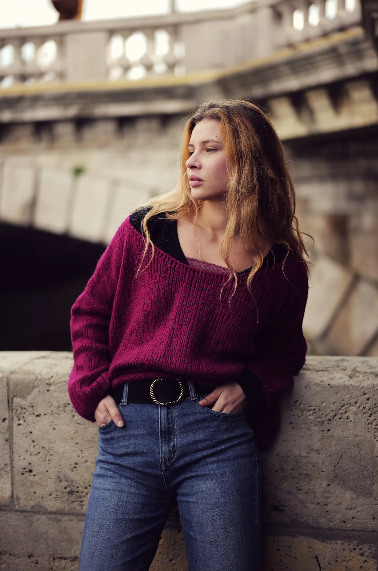 A young woman with long, wavy blonde hair standing by a stone railing outdoors with a bridge in the background, wearing a purple sweater and blue jeans.
