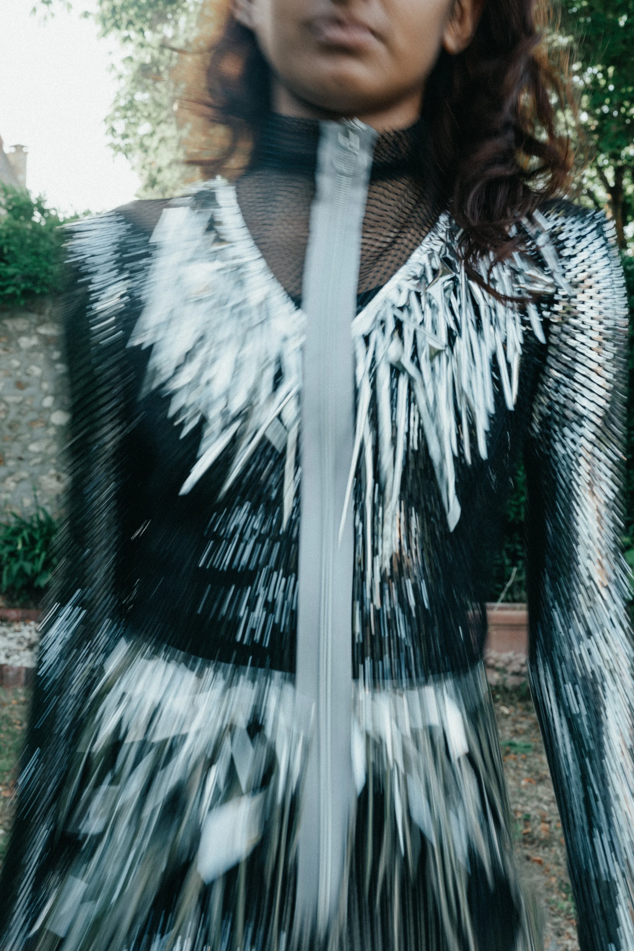 A woman in a futuristic, metallic black and silver jacket with elaborate, spiky shoulder details standing outdoors next to greenery. Raw fashion, up cycling in France. Fashion show in France. 