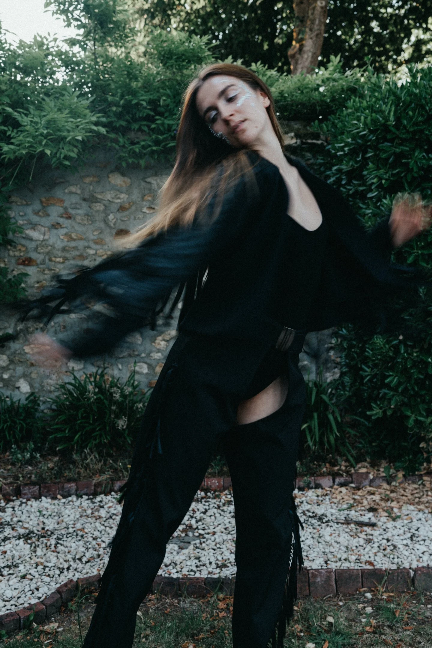 A woman with long hair in a black outfit is posing outdoors among green bushes and trees. She is moving her head and body, creating a dynamic and expressive pose.