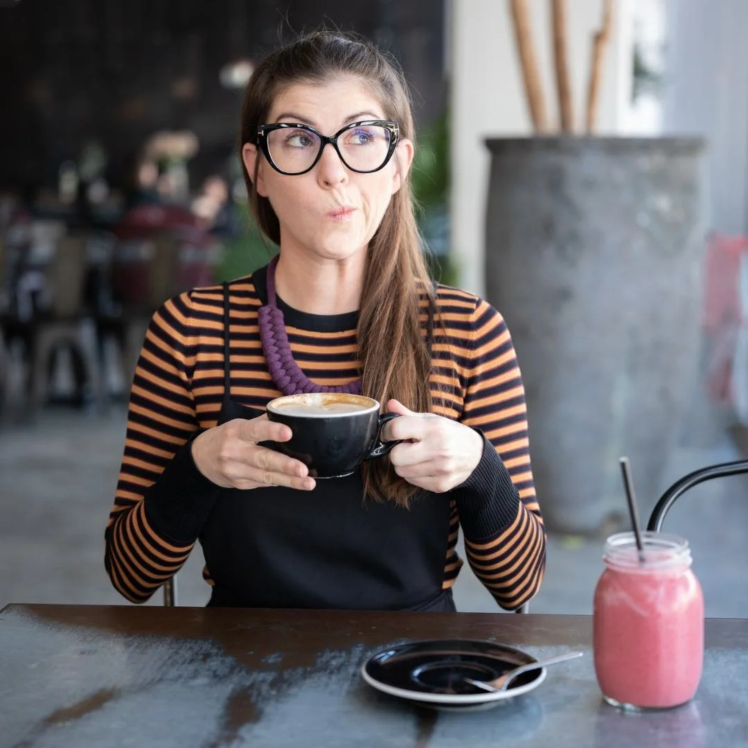 A woman with long brown hair, glasses, and a striped orange and black shirt, sitting at a table in a cafe, holding a black coffee mug and looking to her right with a thoughtful expression, with a pink smoothie with a black straw on the table.