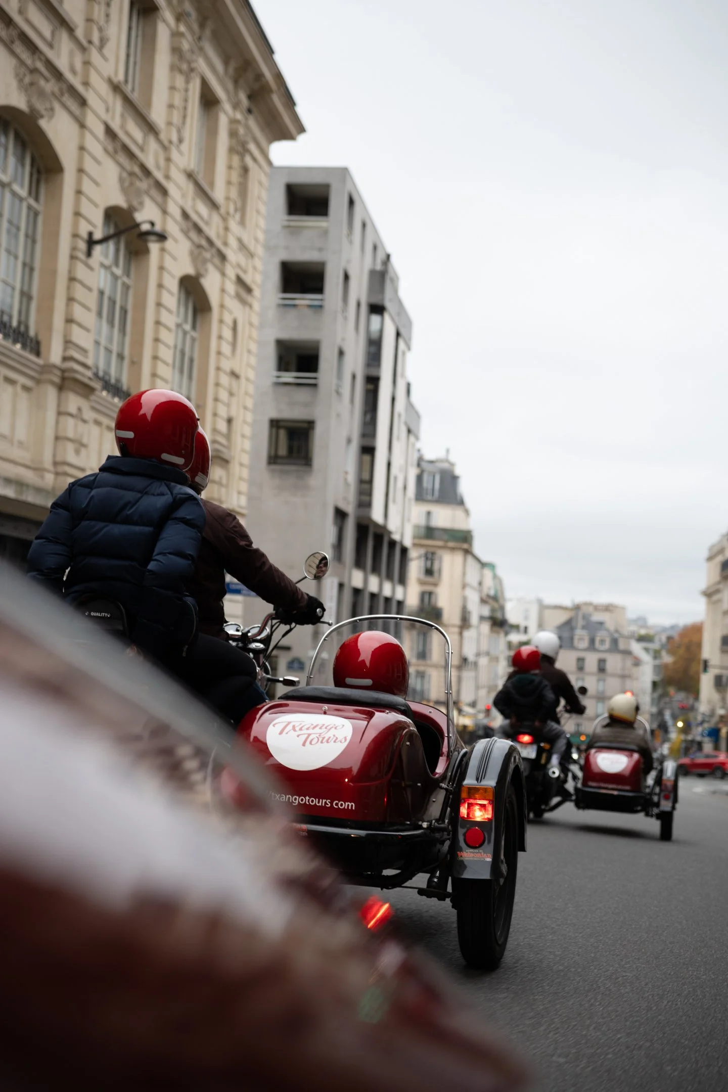 A group of motorcyclists riding together on a city street, many wearing helmets with a red and white star design, and a sidecar labeled 'Trango Tours.' Buildings line the street, and the weather appears overcast.