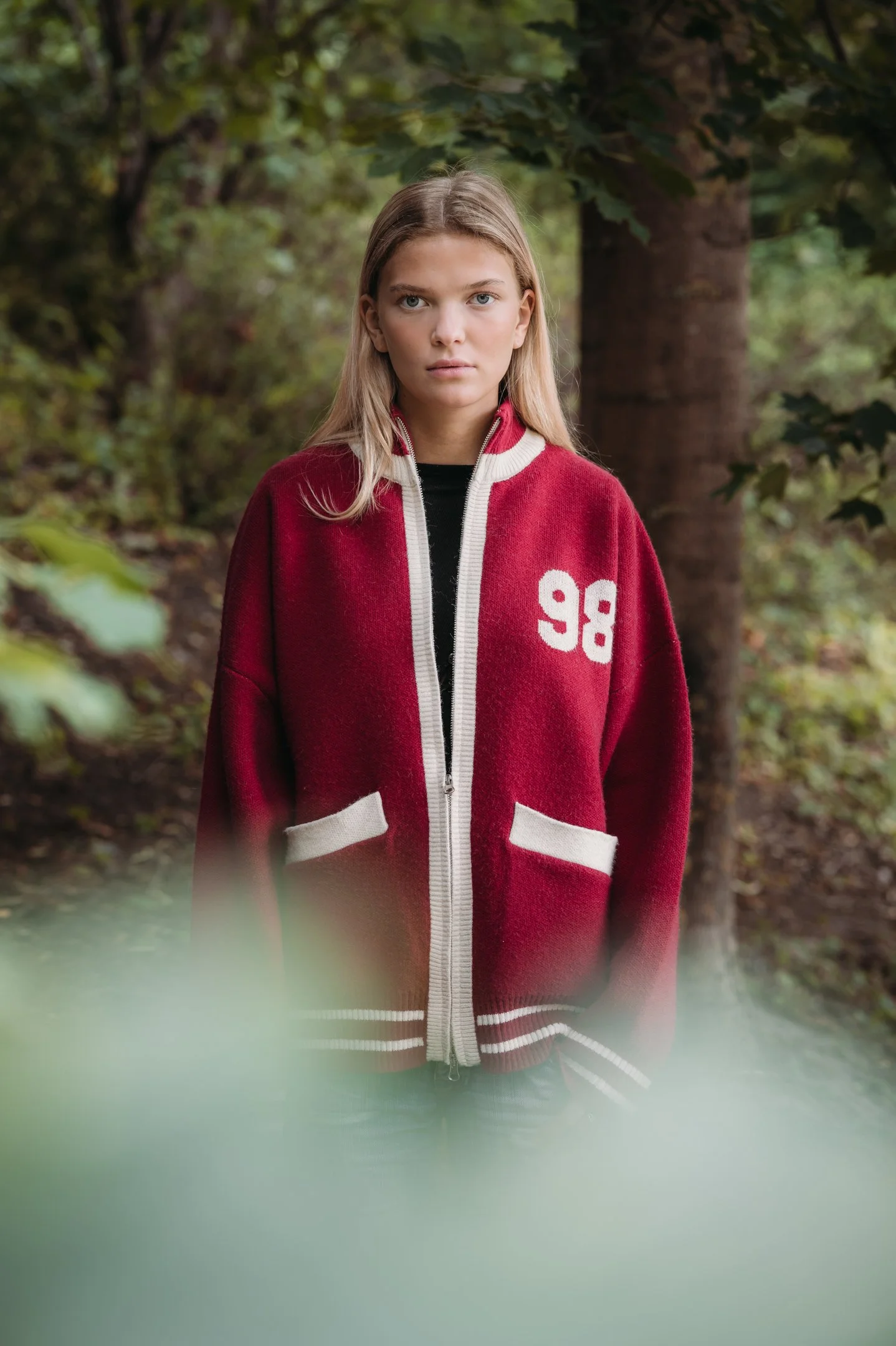 A young woman model with blonde hair and blue eyes standing outdoors in a forest, wearing a red varsity-style jacket in Stockholm Sweden. 