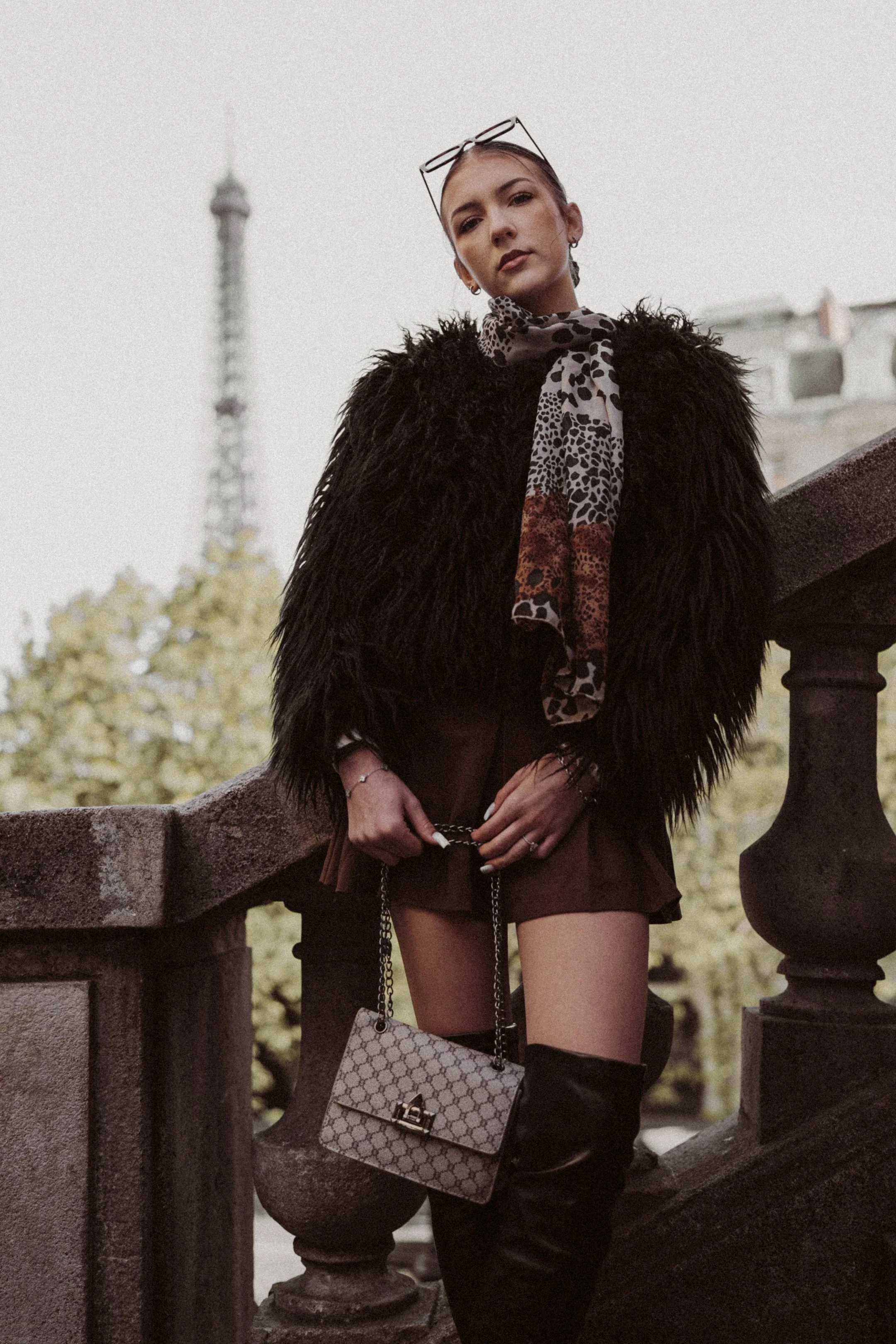 Fashionable woman model standing on a stone staircase with the Eiffel Tower in the background, wearing a black furry coat, patterned scarf, shorts, thigh-high boots, and carrying a designer handbag.