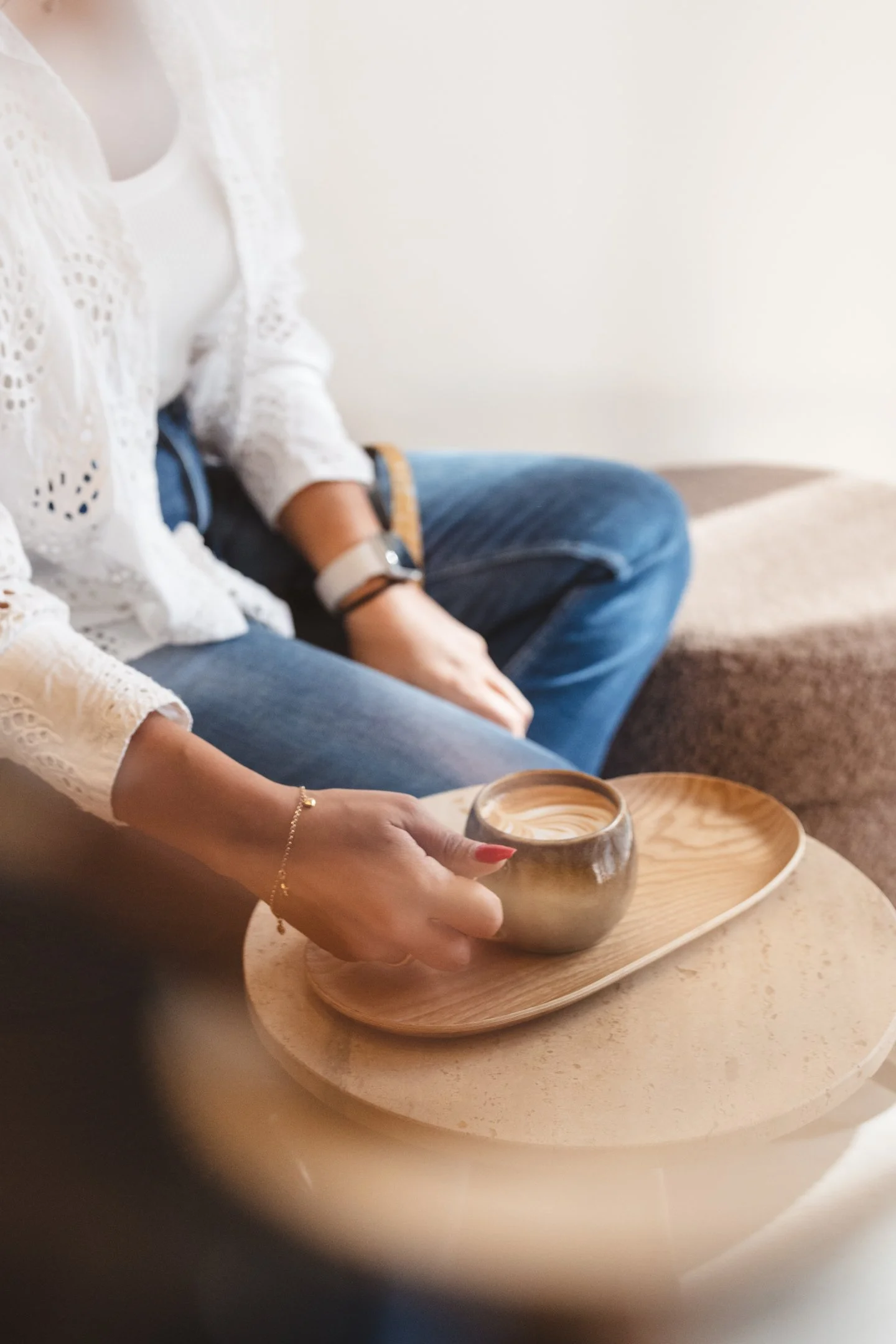 A person wearing a white lace top and blue jeans sits on a beige carpeted floor next to another person. The person is holding a small ceramic cup filled with a hot beverage, placed on a wooden tray. The setting appears cozy and casual, with a minimal