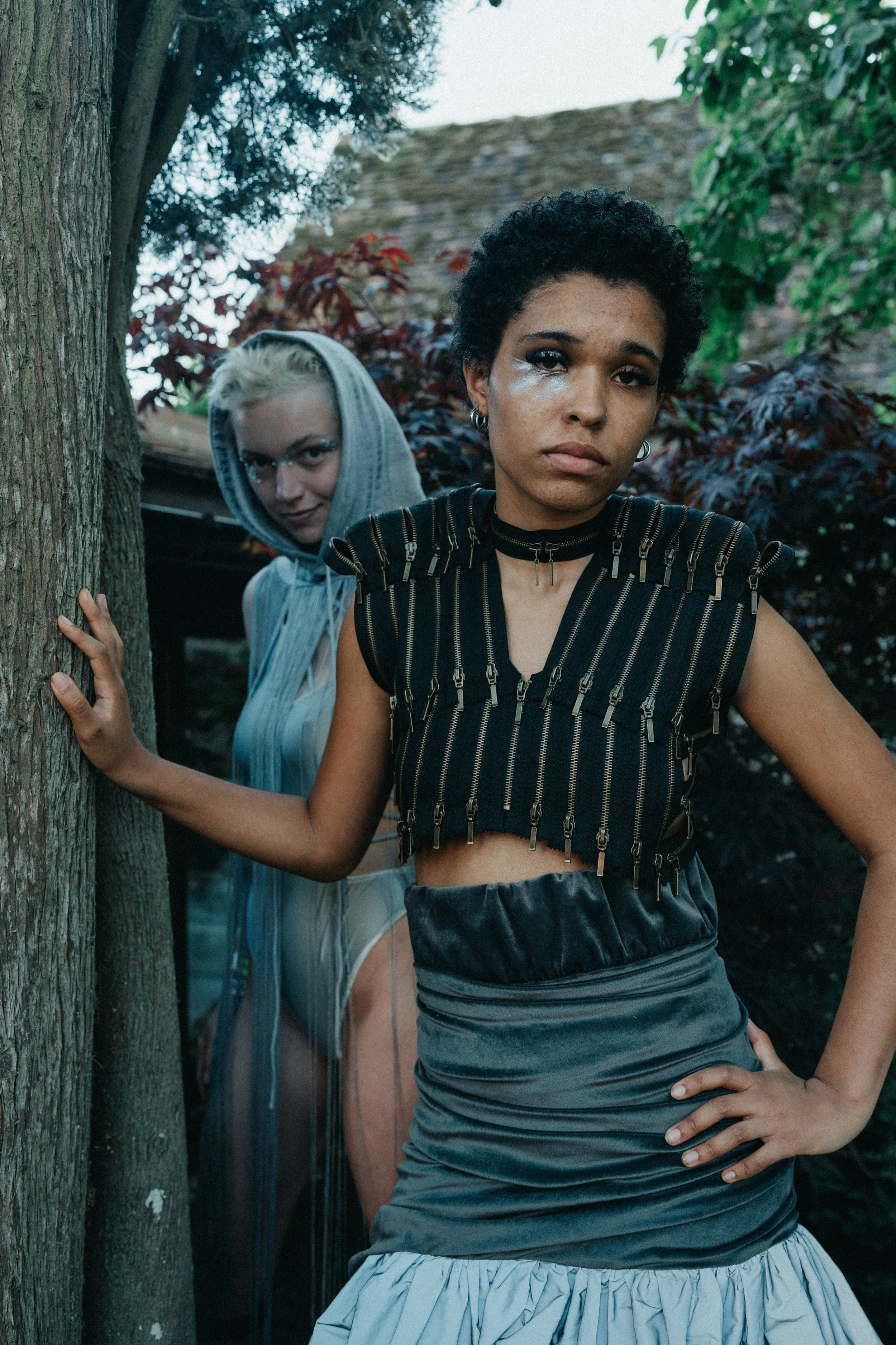 Two women models in France during a photo shoot during a fashion show; hair, a black top with zippers, and a gray skirt, looking serious. The woman in the background has bleach blonde hair, wears glasses, a hooded top, and looks mischievous.