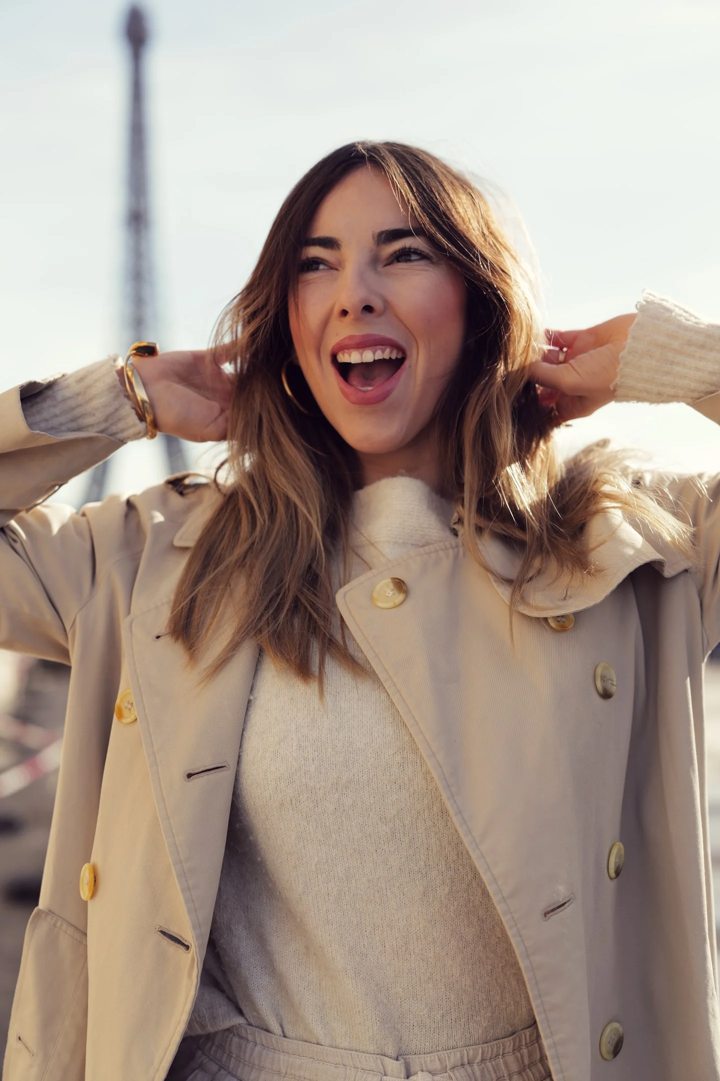 A woman with long brown hair smiling outdoors, wearing a beige trench coat, a cream-colored sweater, and gold hoop earrings, with her hands behind her head, in a cityscape setting.