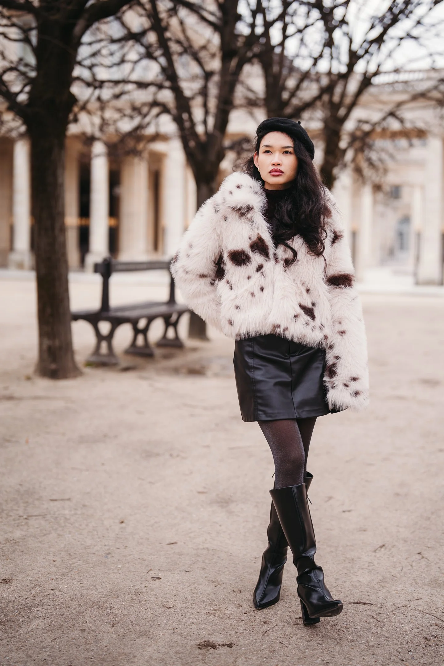 A woman with long dark hair wearing a white fur coat with brown spots, black hat, black leather skirt, black tights, and high-heeled black boots stands outdoors in front of leafless trees and a building with columns.