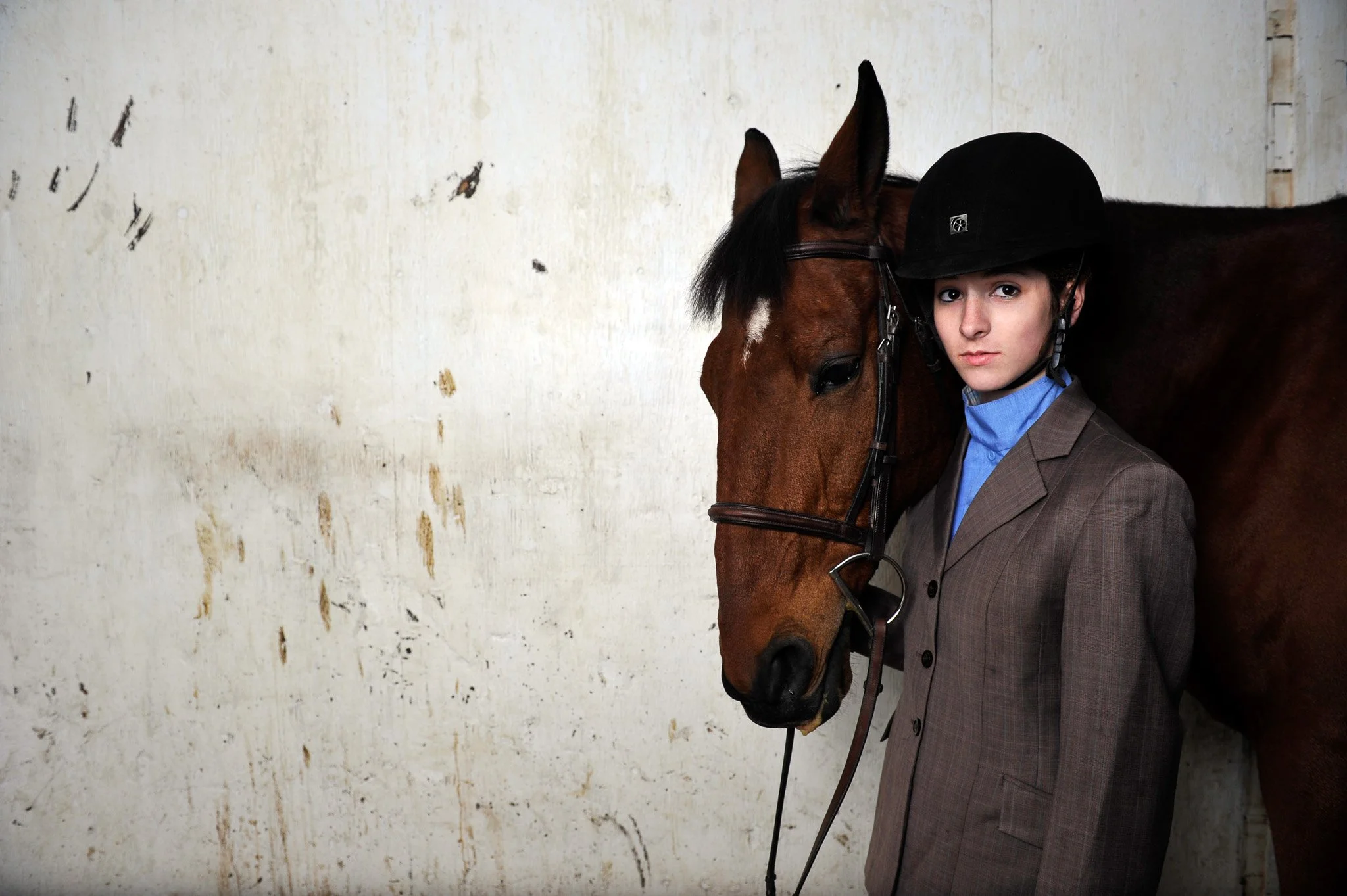 Young equestrian in a suit and riding helmet standing close to a brown horse against a worn white wall.