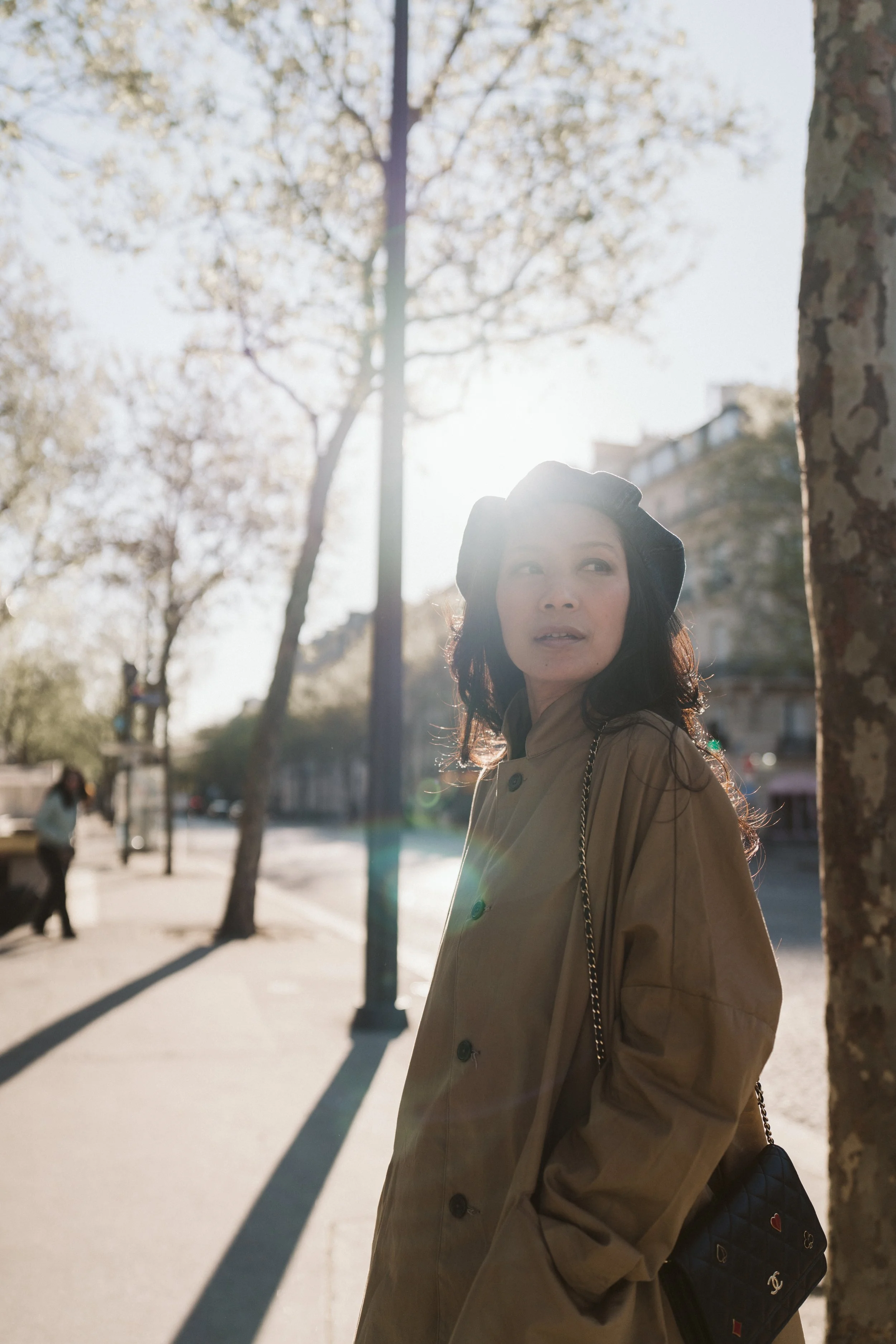 Woman with dark hair wearing a beige trench coat and a black beret stands on a Parisian sidewalk in Paris 8e with trees and buildings in the background, backlit by sunlight.