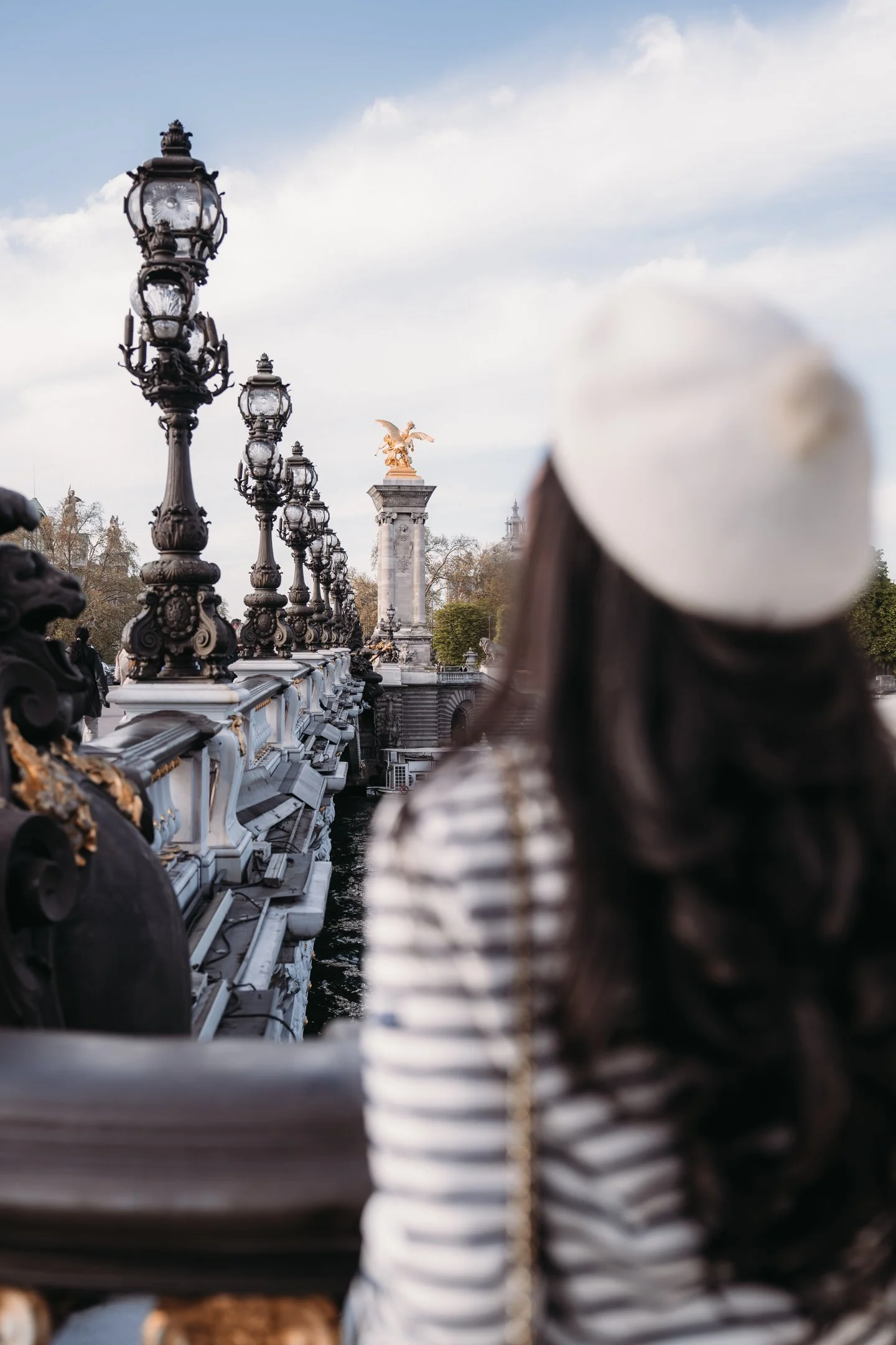 View of ornate street lamps along a bridge, with a gilded angel statue on a stone pedestal in the distance, under a partly cloudy sky.