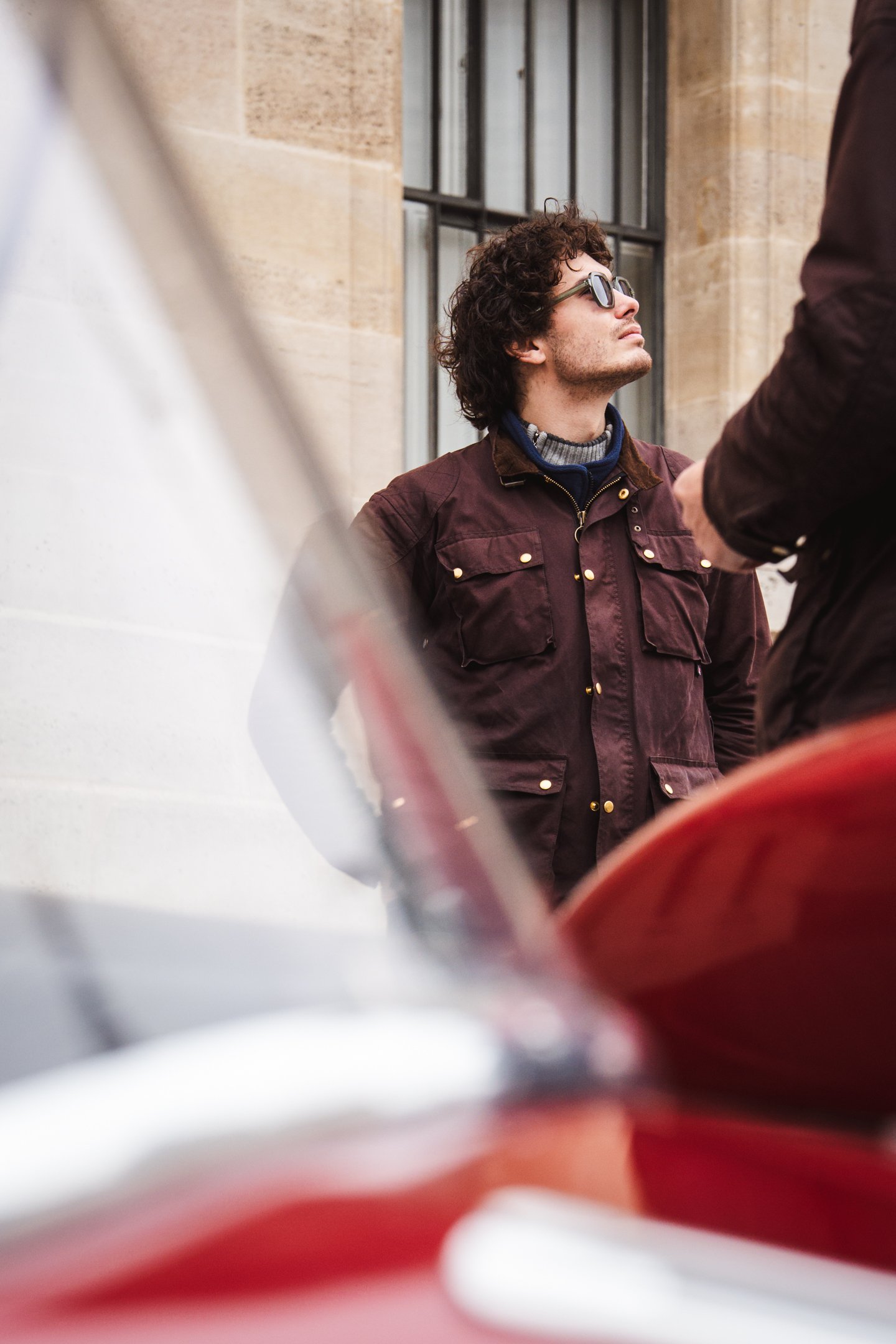 A young man with curly dark hair and sunglasses looking upward, standing outside near a building with beige stone walls and windows. There are blurred objects in the foreground.