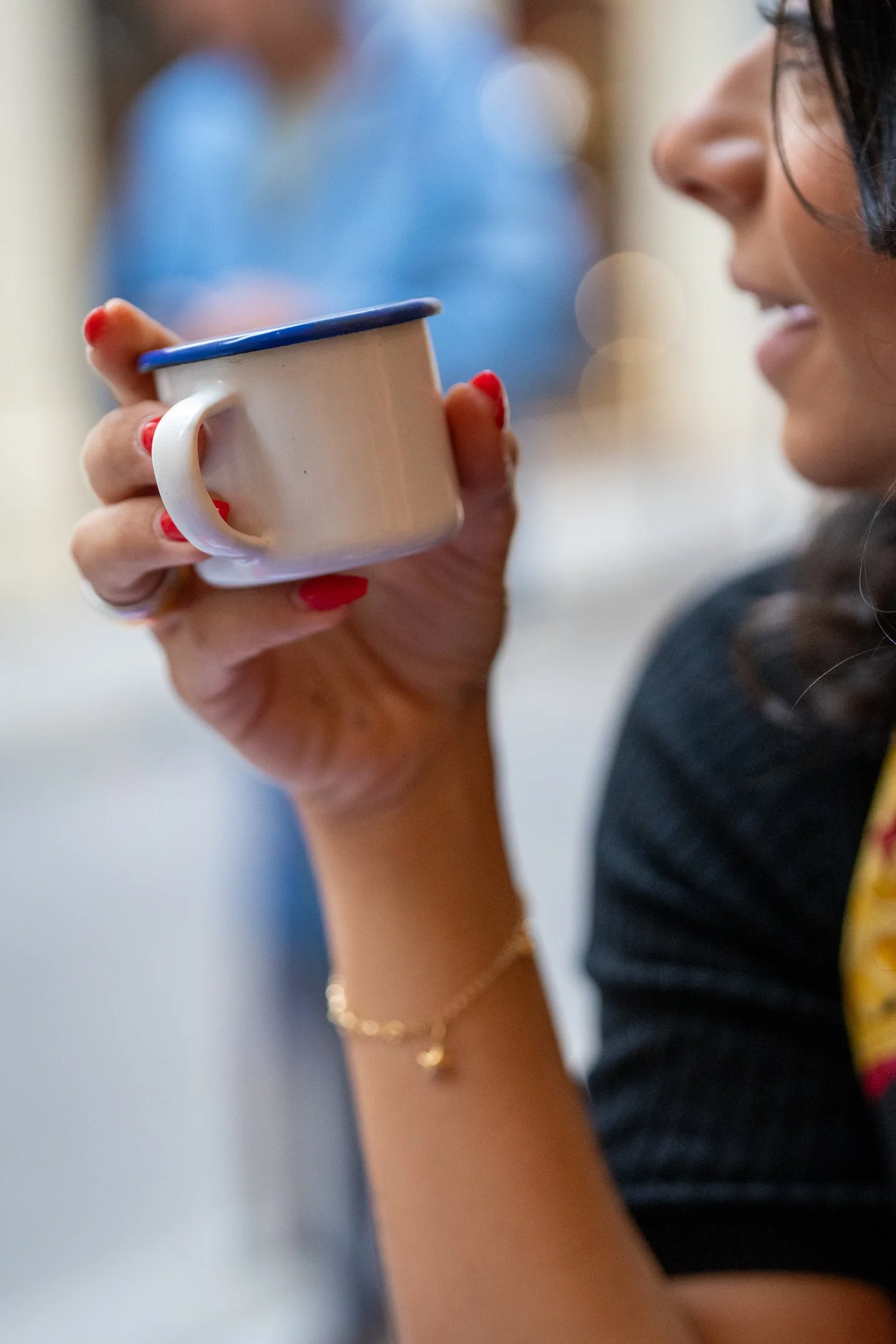 A woman holding a white mug with blue trim, wearing a gold bracelet, and a gold necklace. Her face is turned slightly, and she is smiling. The background is blurred, with a person in a blue shirt visible.