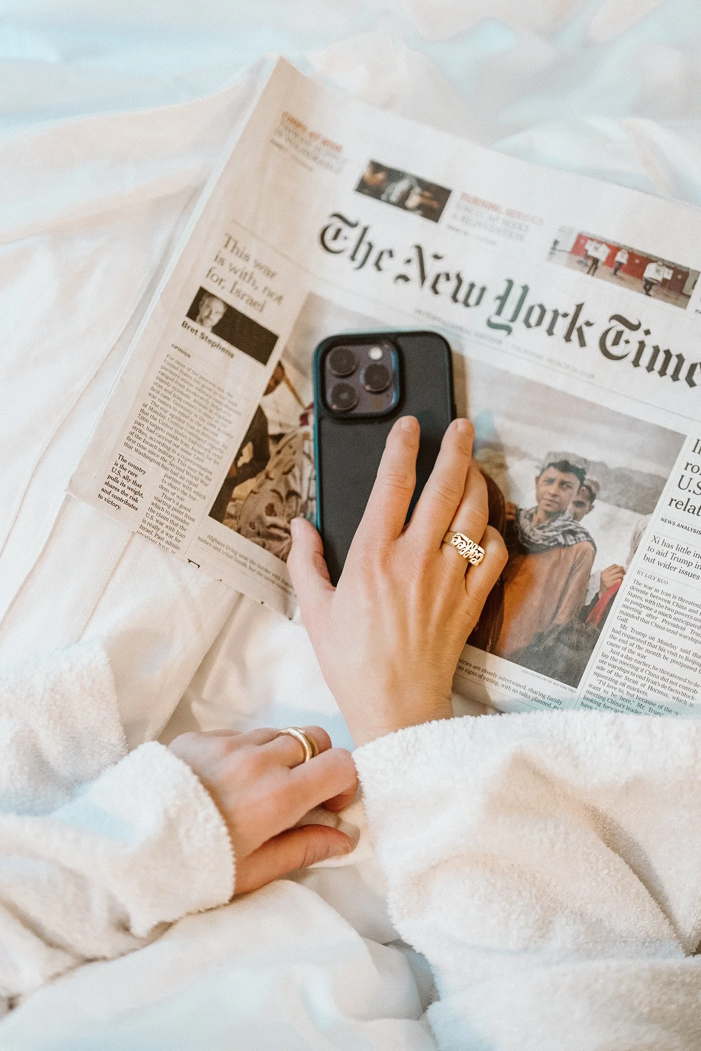 A person wearing a white sweater holding a smartphone above a open newspaper on a light-colored surface.