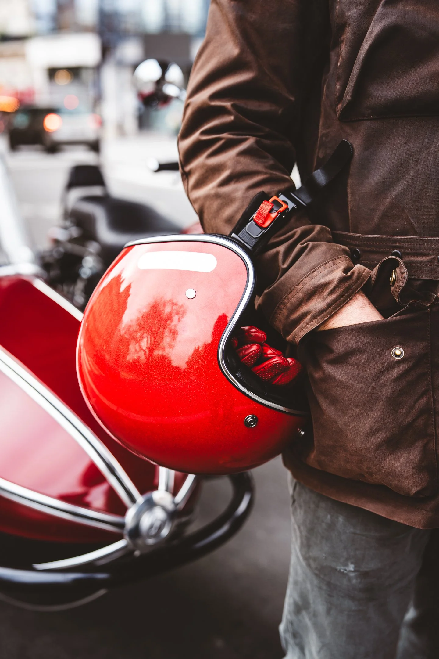A person wearing a brown jacket and gray pants, securing a red motorcycle helmet with a black strap that has a red buckle.