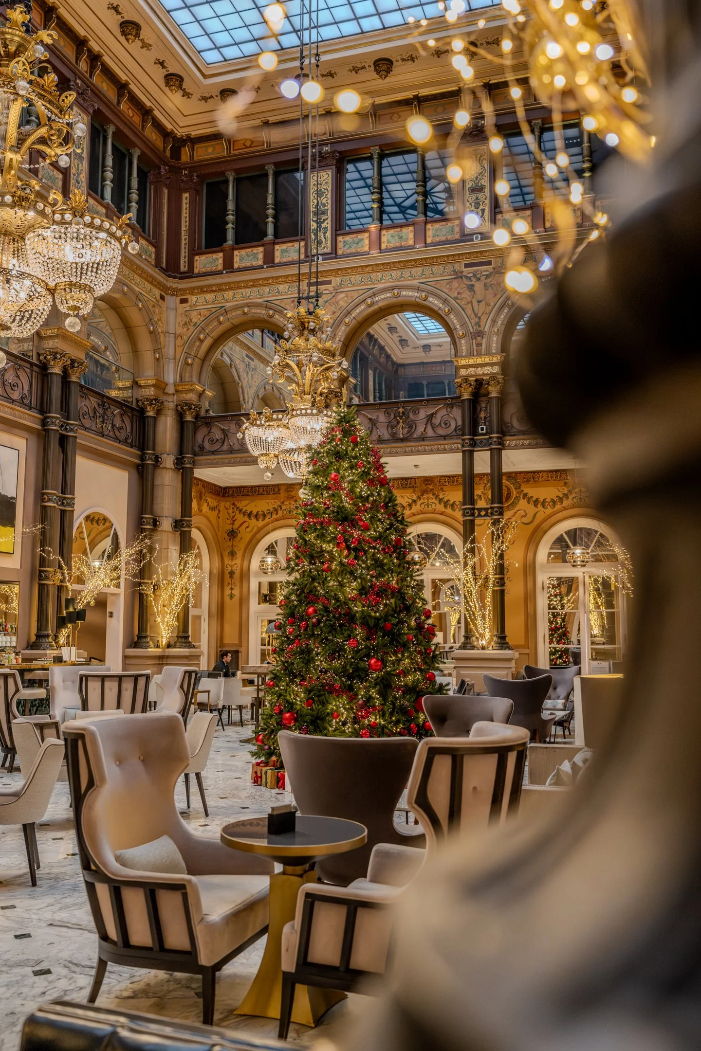 A decorated Christmas tree with red ornaments and gifts underneath is in the center of an elegant, ornate room with chandeliers, arches, and detailed decor. The room has large windows and a glass ceiling, with seating and tables arranged around the t