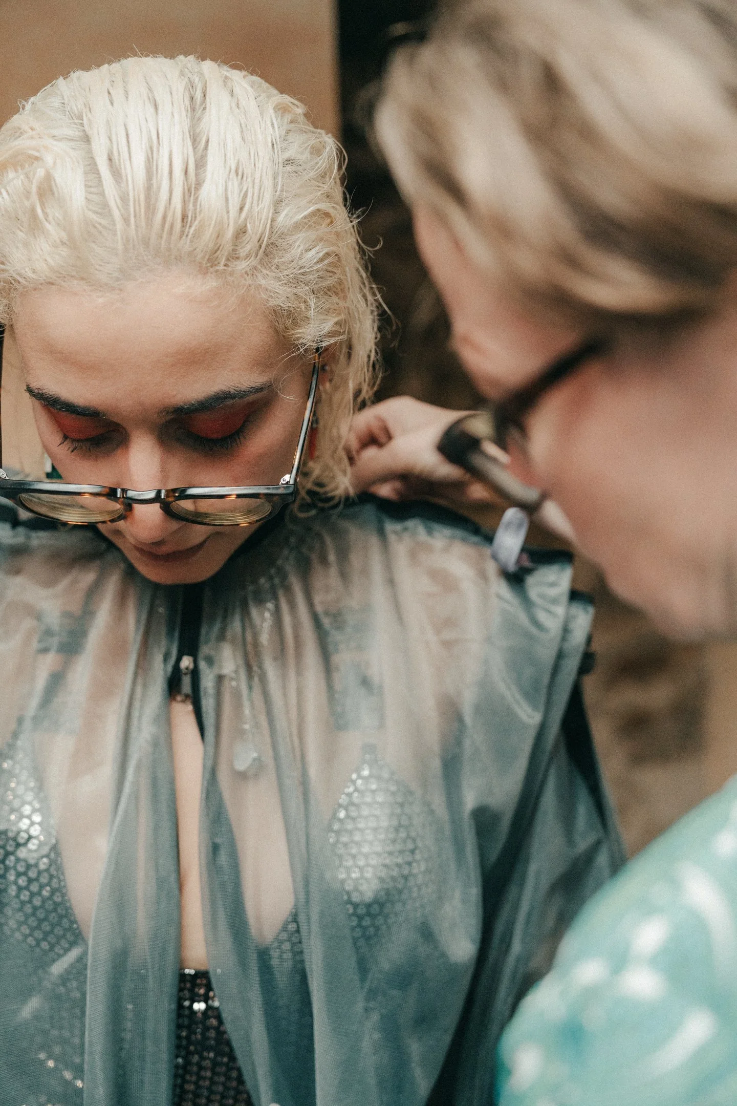 A person with platinum blonde curly hair, wearing dark glasses, getting a tattoo on their shoulder by a tattoo artist.