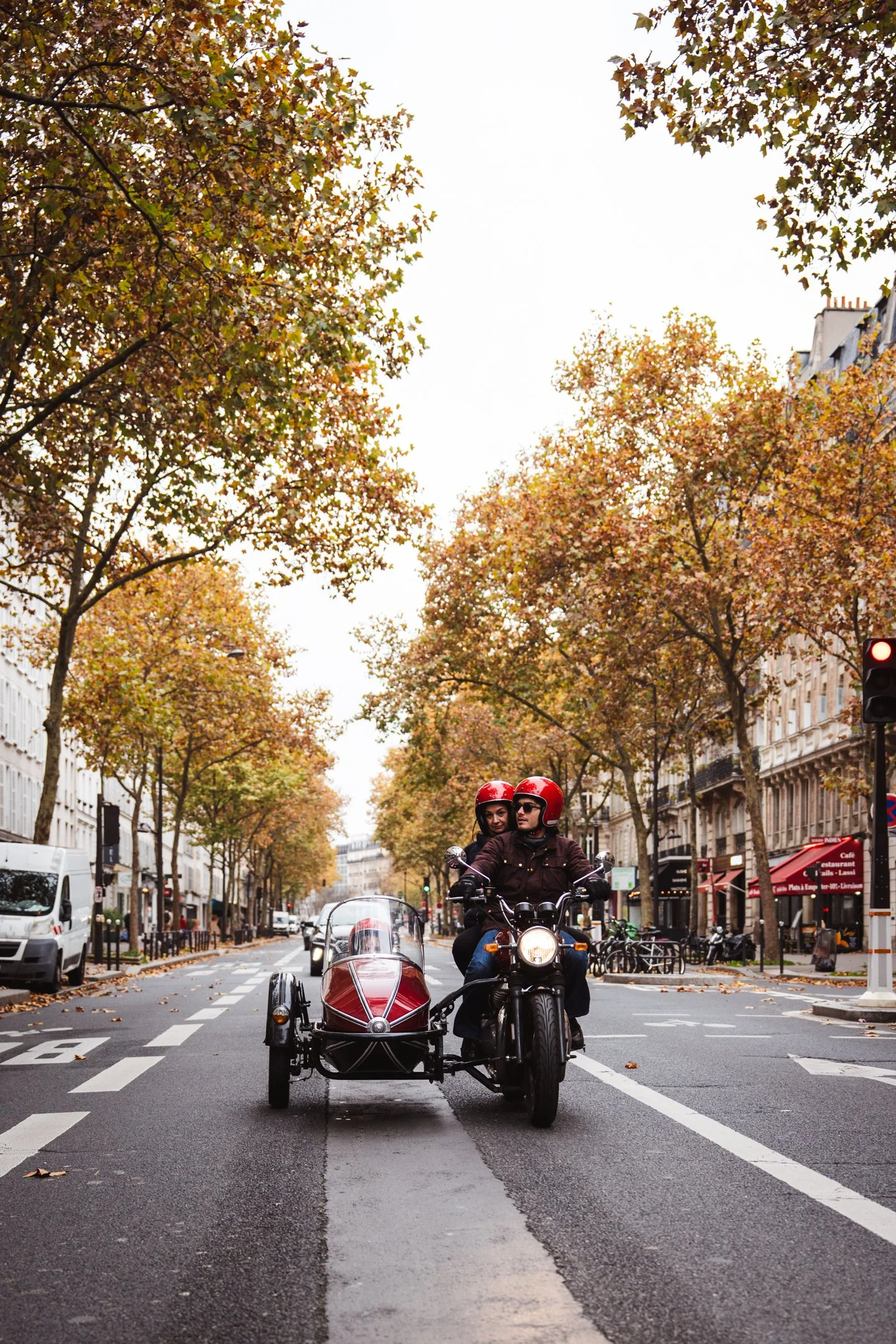 A man and woman riding a motorcycle on a city street, with a sidecar, surrounded by parked cars, trees with autumn leaves, and buildings. The woman is sitting behind the man, both wearing helmets.