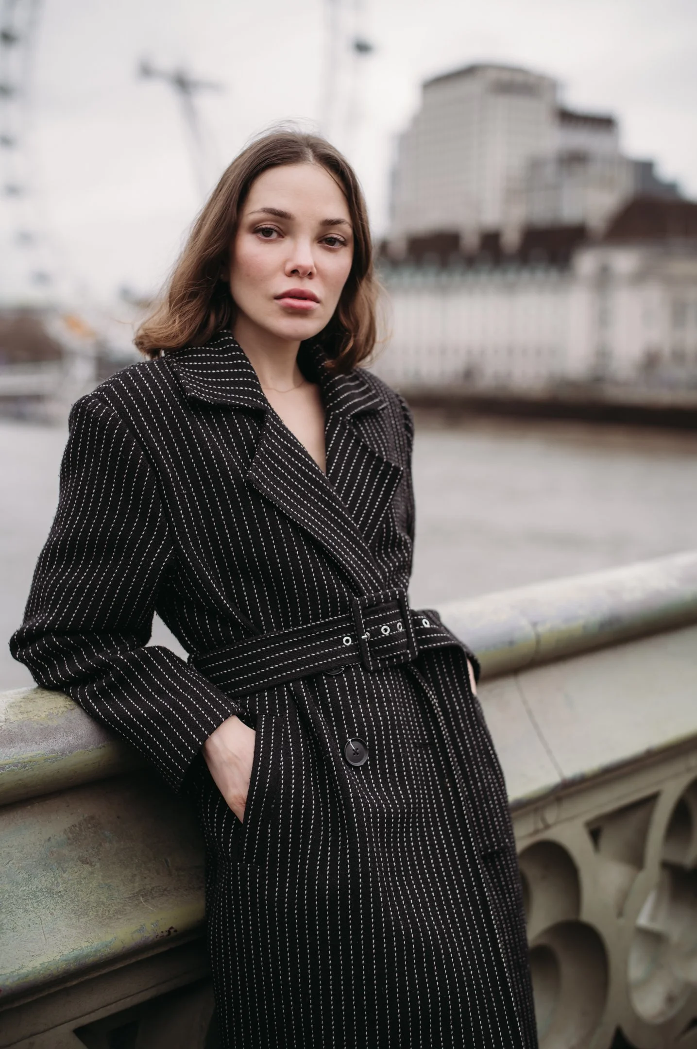 A young woman with shoulder-length brown hair, wearing a black pinstripe coat, stands outdoors leaning against a stone railing with her right hand in her coat pocket, with urban buildings and a Ferris wheel blurred in the background.