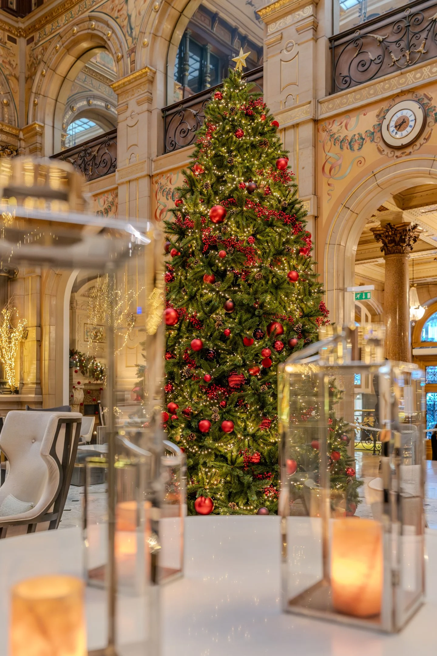 Christmas tree decorated with red and gold baubles, ribbons, and fairy lights in an ornate, elegant hall with large arches, chandeliers, and intricate wall decor.