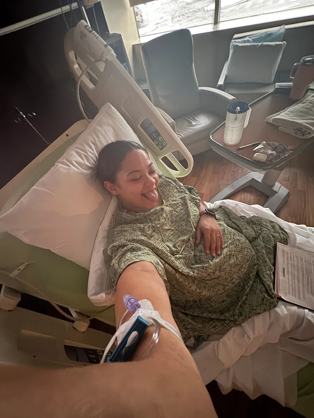 A woman lying in a hospital bed with an IV in her arm, smiling and sticking out her tongue. She is wearing hospital clothing, and the room includes a chair, a tray table with a drink, and medical equipment.
